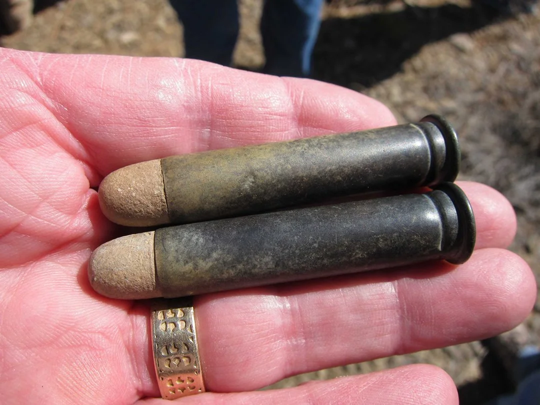 A person's hand holding two old, weathered artillery shells with black rings on one end and a ring on the finger. The shells are tarnished and appear rusty.