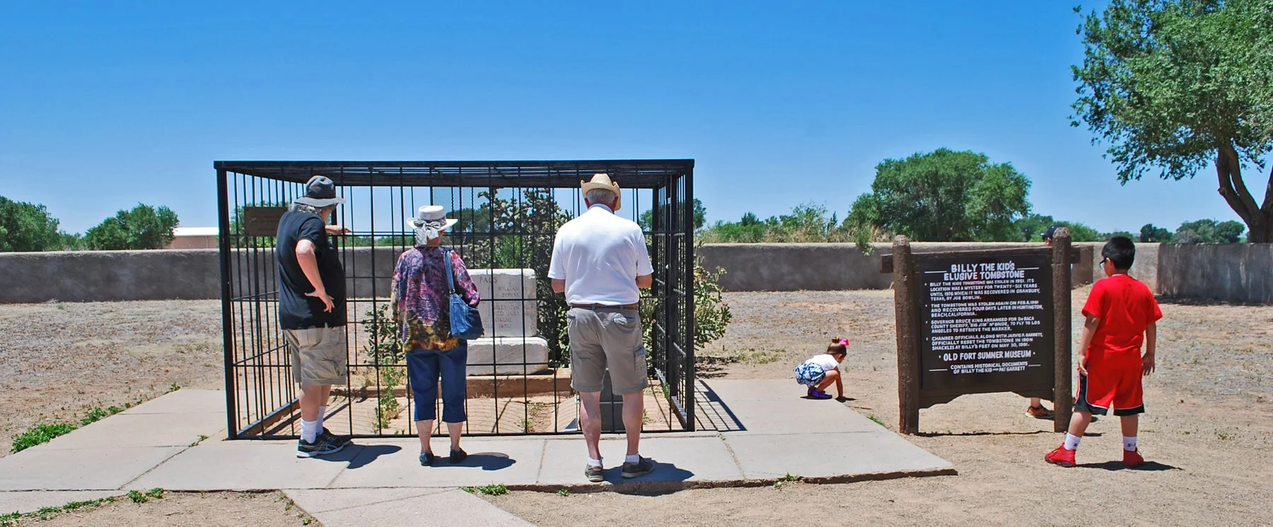 Group of five people, including children, visiting the Billy the Kid's Elusive Tombstone exhibit at a museum, with a large sign explaining its history, outdoors on a sunny day.