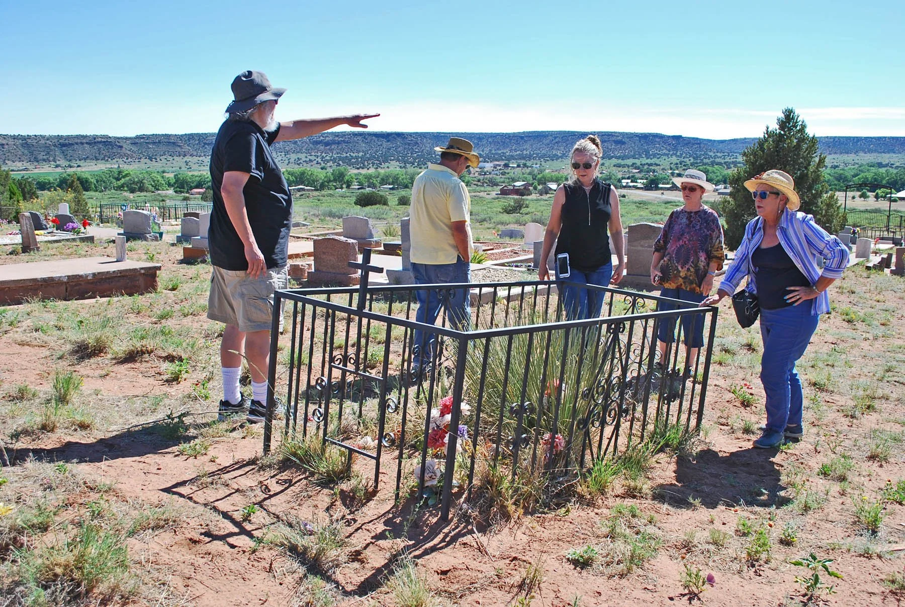 A group of six people gathered around a grave site in a rural cemetery on a sunny day. They are dressed casually, with some wearing hats and sunglasses, and appear to be engaged in a discussion or ceremony.