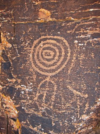 Ancient rock petroglyph of a spiral pattern connected to a circle and lines, etched into a dark sandstone surface.