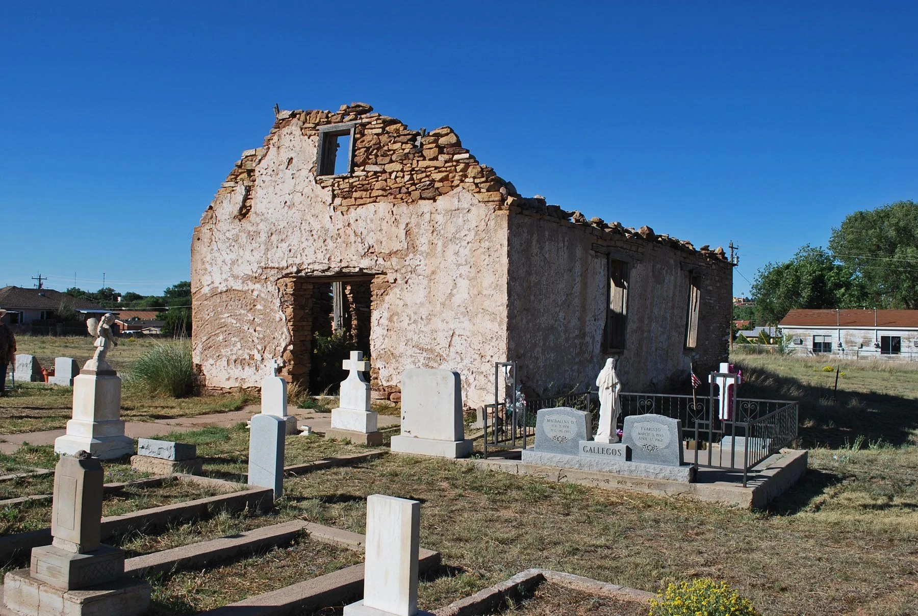 A dilapidated stone and plaster church in a cemetery, with several graves marked by tombstones and small statues. The church has missing sections of its roof and walls, with a clear blue sky in the background.