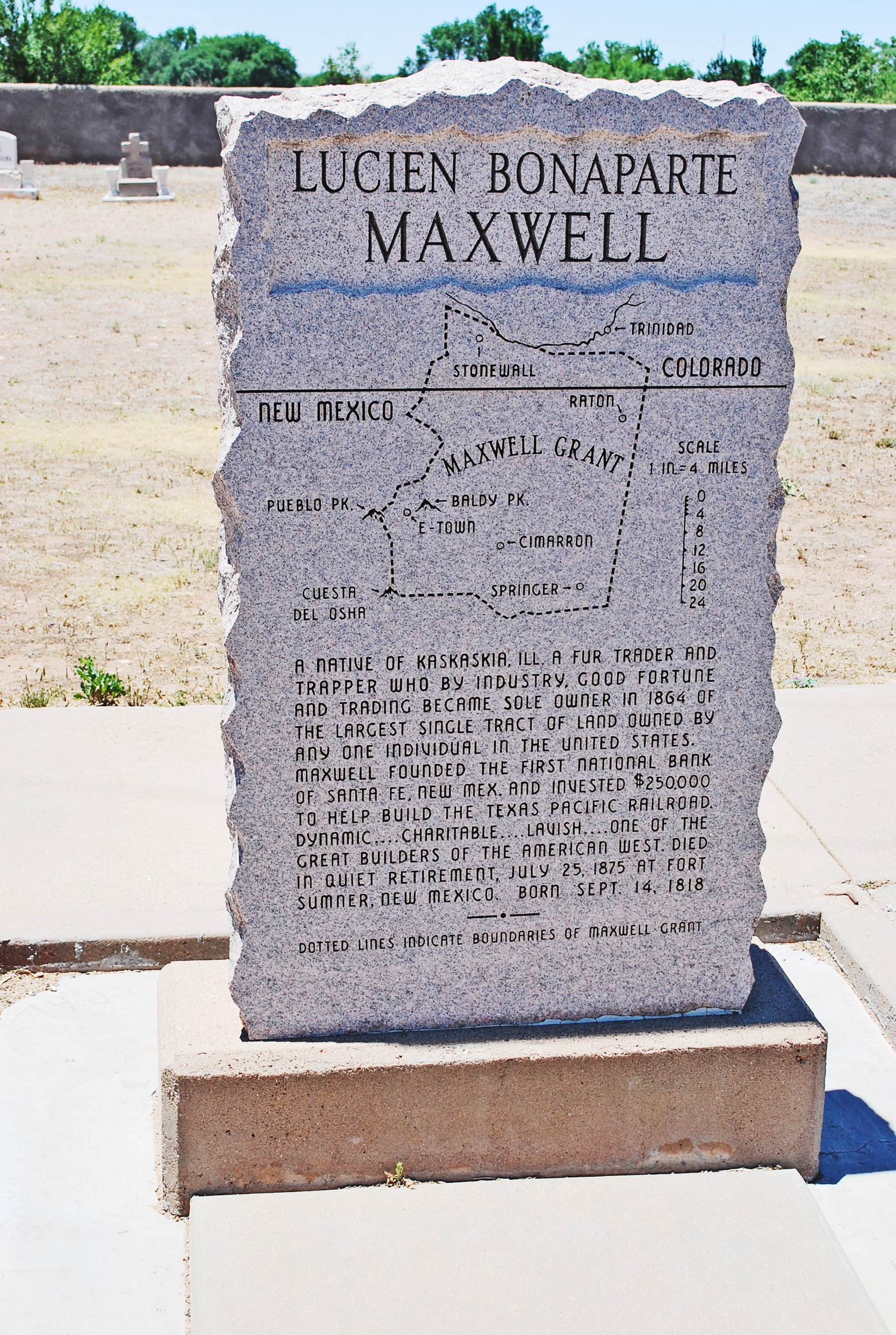 A granite monument with a map and text about Maxwell Grant, a fur trader and landowner born in 1818, located outdoors with a clear sky and trees in the background.