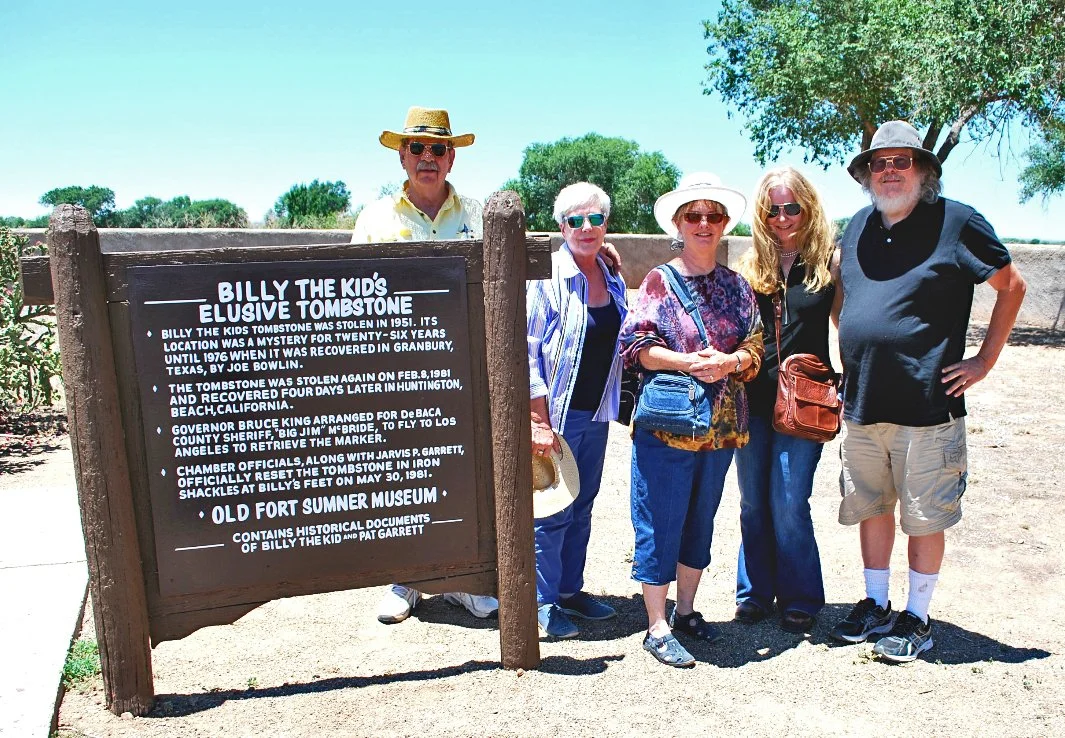 Group of five people standing outdoors next to a historical marker about Billy the Kid's Tombstone, with a tree and clear blue sky in the background.