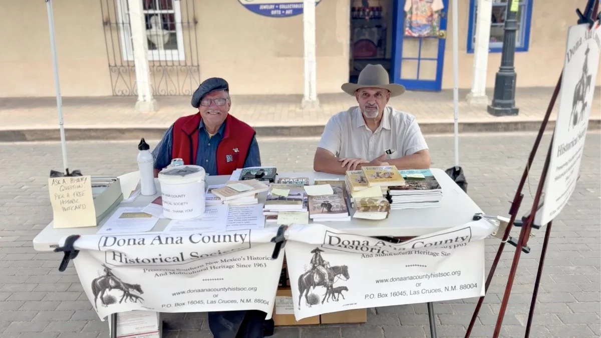 Two men sitting at a table with historical books and materials, representing the Dona Ana County Historical Society, on a city sidewalk.
