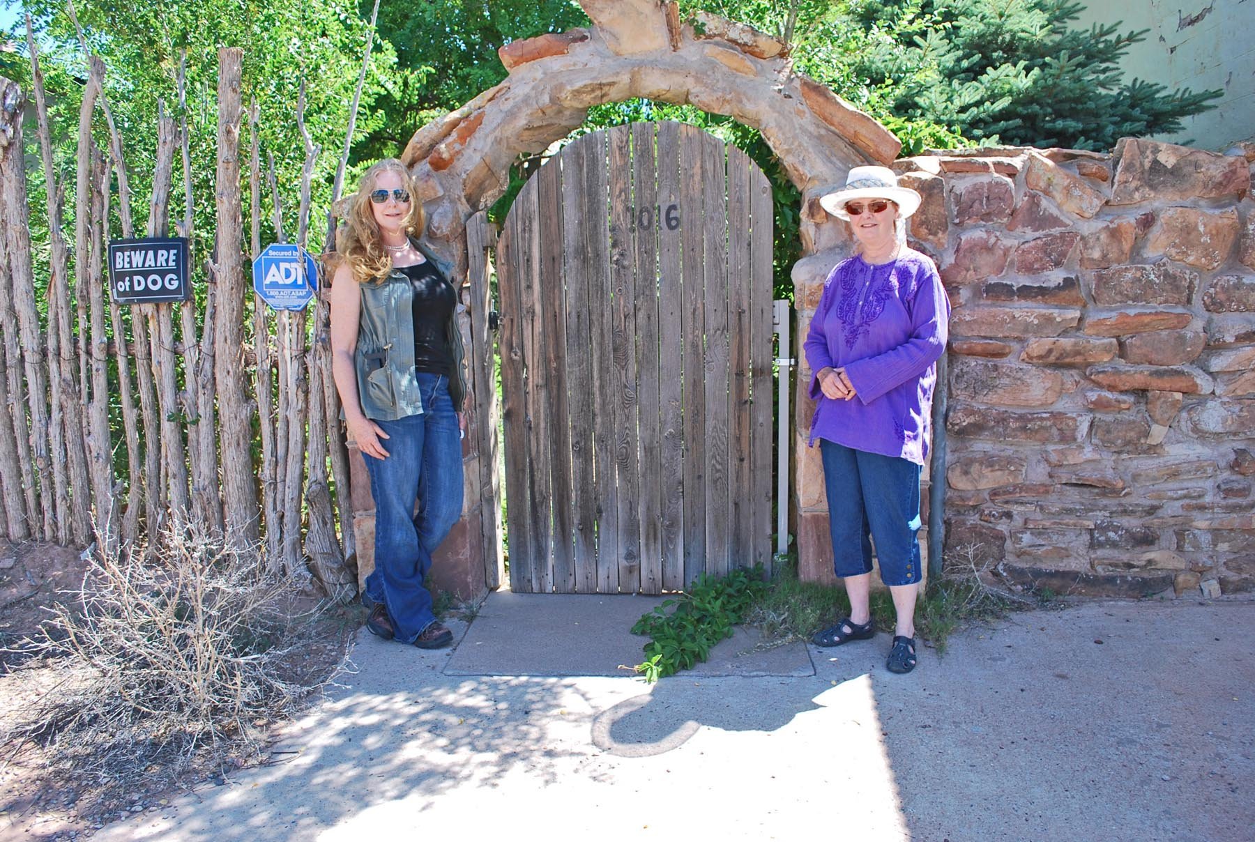 Two women standing in front of a wooden gate with a stone arch and decoration, behind a rustic wooden fence with signs warning of a dog and security camera, surrounded by greenery and trees.