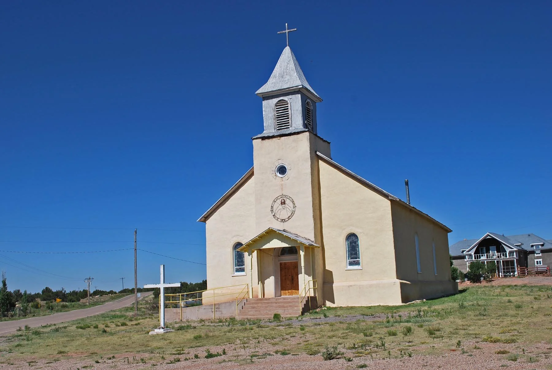 A small beige church with a wooden door, a cross at the top of the steeple, and a clock on the front, set against a bright blue sky with some clouds and nearby residential houses.