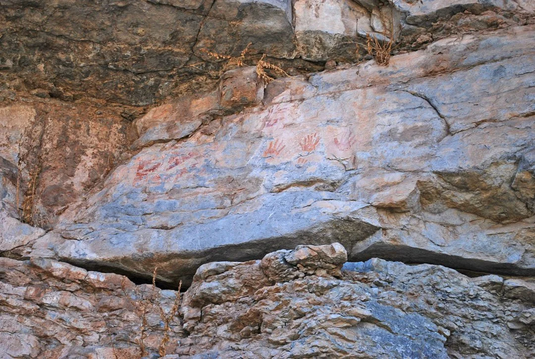 Ancient rock wall with red and orange handprint and figure petroglyphs.