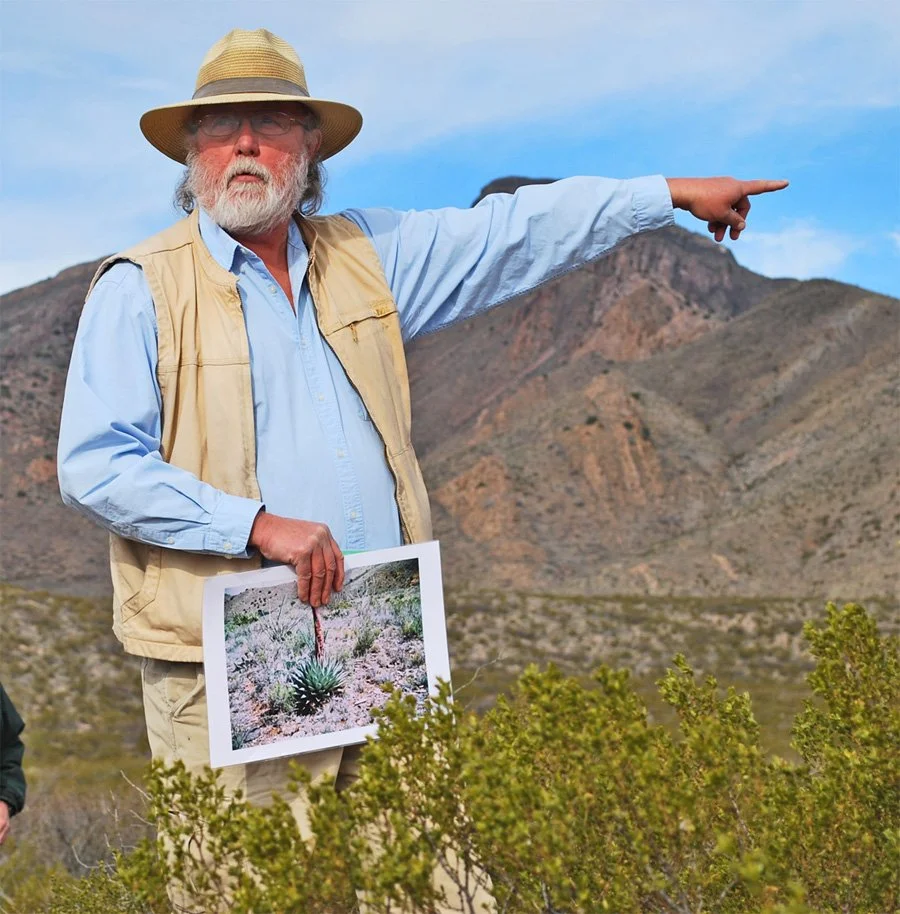 An older man with a beard, wearing a straw hat, glasses, a light blue shirt, and a beige vest, points to the right while holding a photo of a desert plant. The background features mountains and a partly cloudy sky.