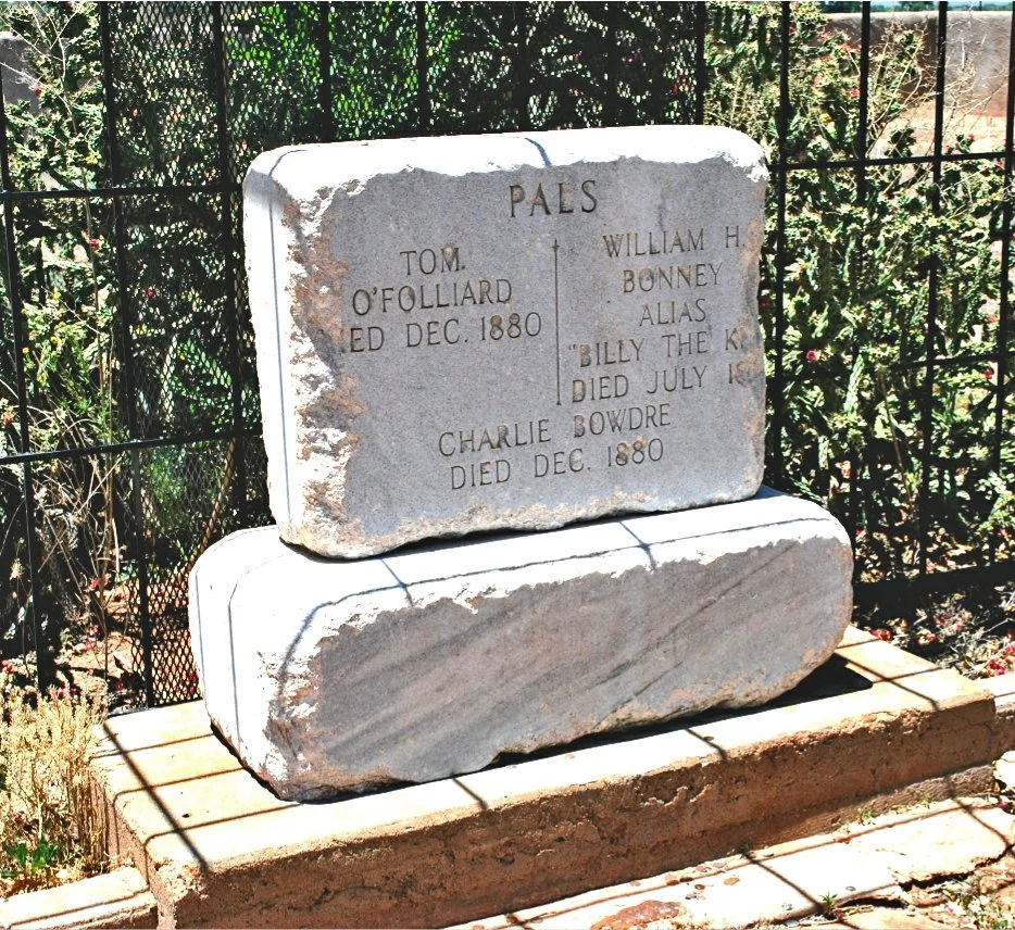 A stone memorial with engraved names and dates, set on a brick base, surrounded by a metal fence and greenery.