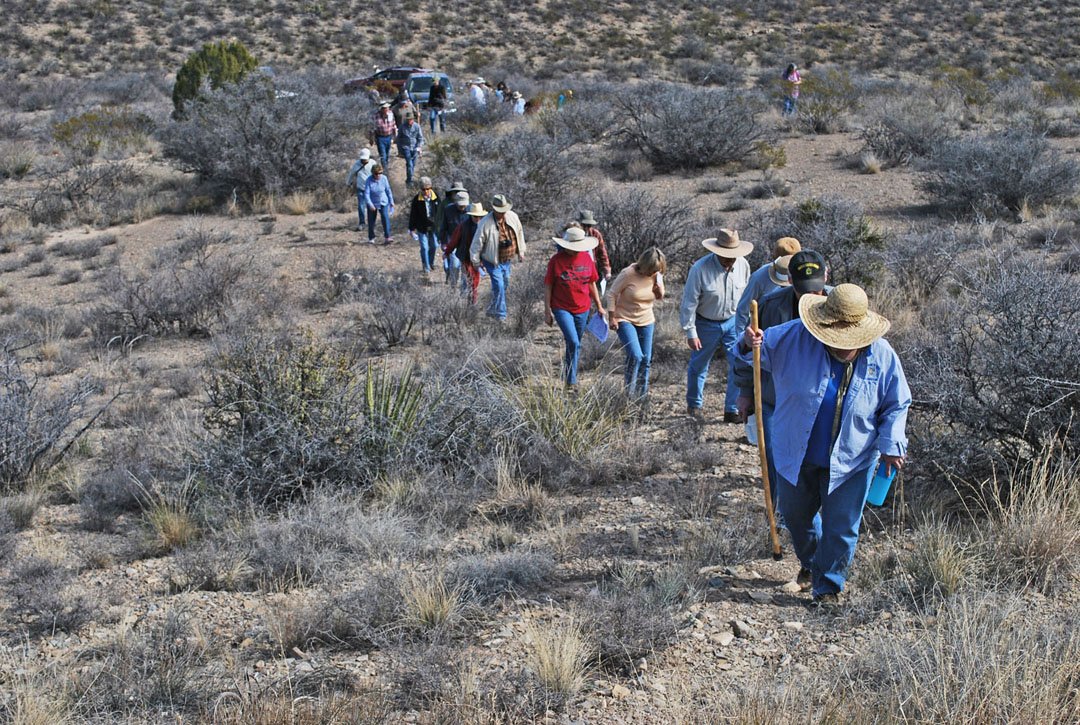 A group of people hiking up a trail in a desert landscape, wearing hats and casual outdoor clothing.