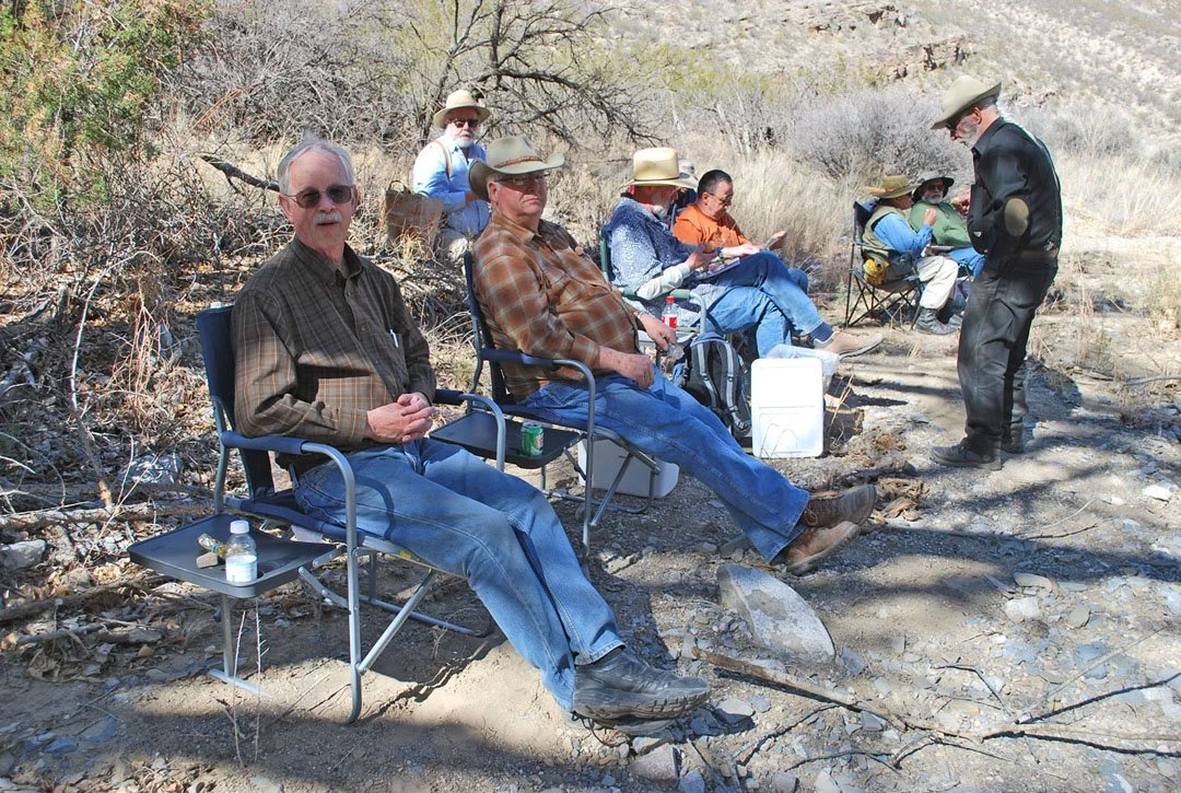 Group of people sitting on chairs outdoors in a rocky, dry landscape with sparse bushes, some wearing hats and sunglasses.