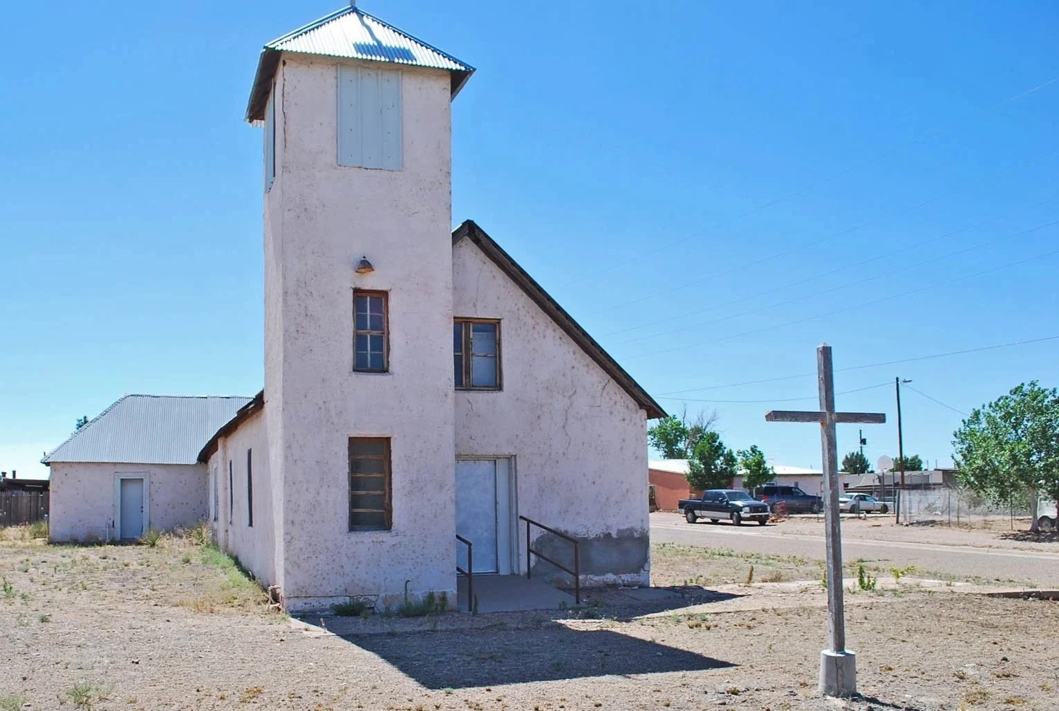 An old white church with a tall tower, a small side building, and a wooden cross outside. The church is in a desert area with parked cars and a clear blue sky.