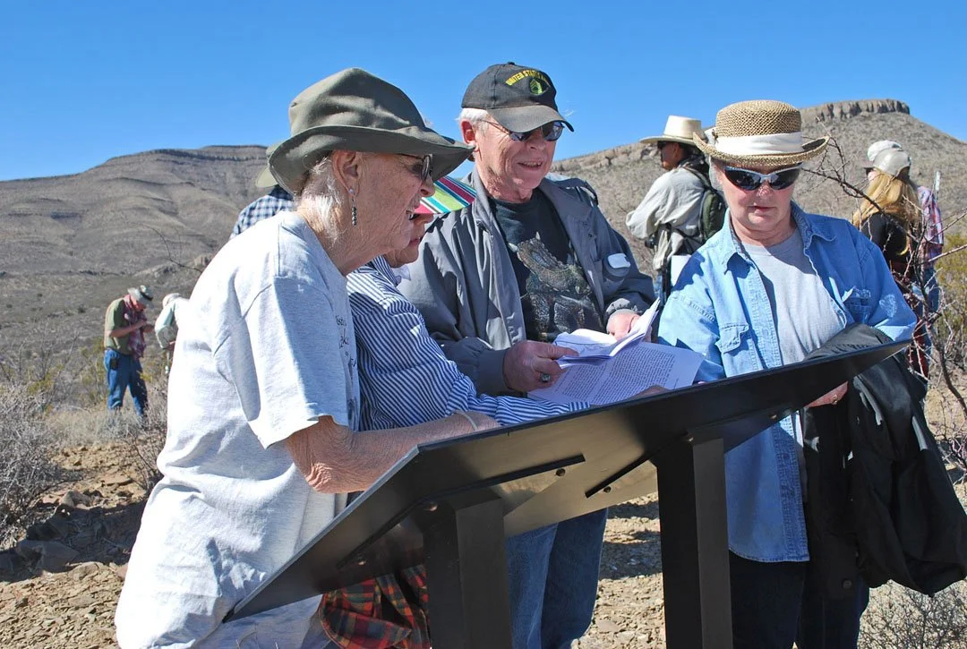 Group of older adults examining a sign or map outdoors in a desert landscape under clear blue sky, wearing hats and sunglasses.