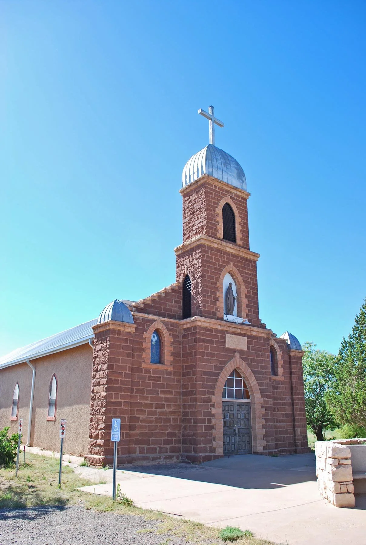 A red brick church with a steeple topped by a cross, set against a clear blue sky. The church has arched windows and a large wooden door, with small metal domes on the roof and a religious statue in an alcove above the main entrance.
