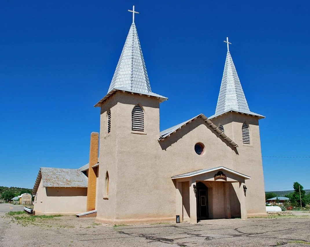 A beige church with two tall steeples topped with white crosses, a small entrance porch, and a bright blue sky in the background.