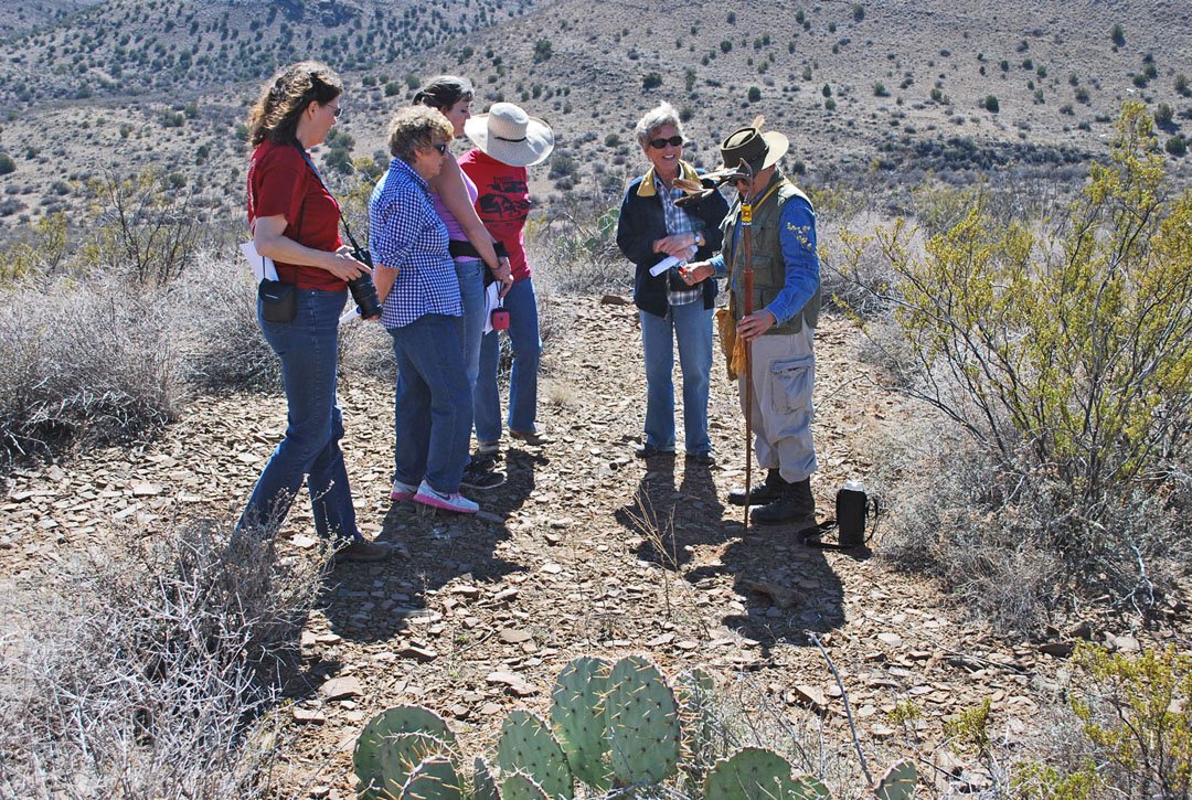 A group of six people on a desert trail listening to a park ranger. The surroundings are arid with cacti and dry bushes.