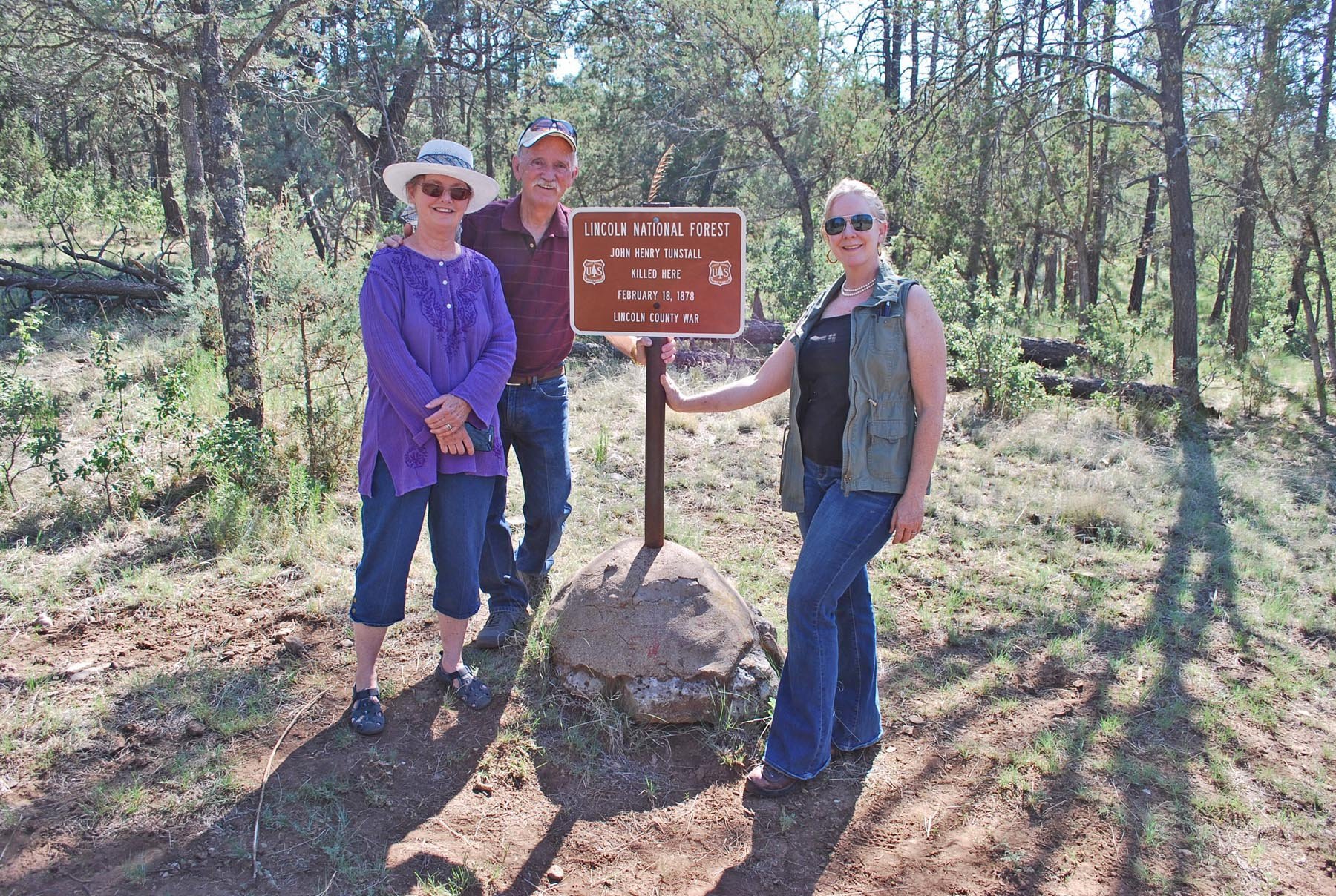 Three smiling people standing around a historical marker sign at Lincoln National Forest, surrounded by trees and greenery, on a sunny day.