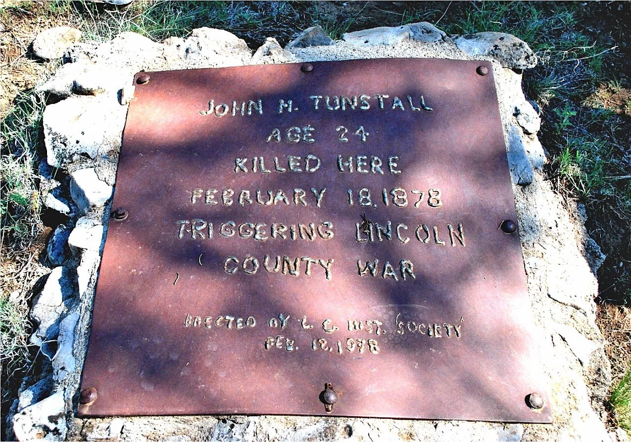 A metal memorial plaque dedicated to John M. Tunstall, who was killed here at age 24 during the Lincoln County War on February 18, 1878. The plaque is mounted on rocks and surrounded by grass.
