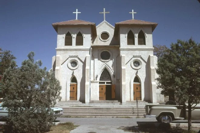 Front view of a white church with three crosses on the roof, arched windows, and wooden doors, with trees and parked cars in front.