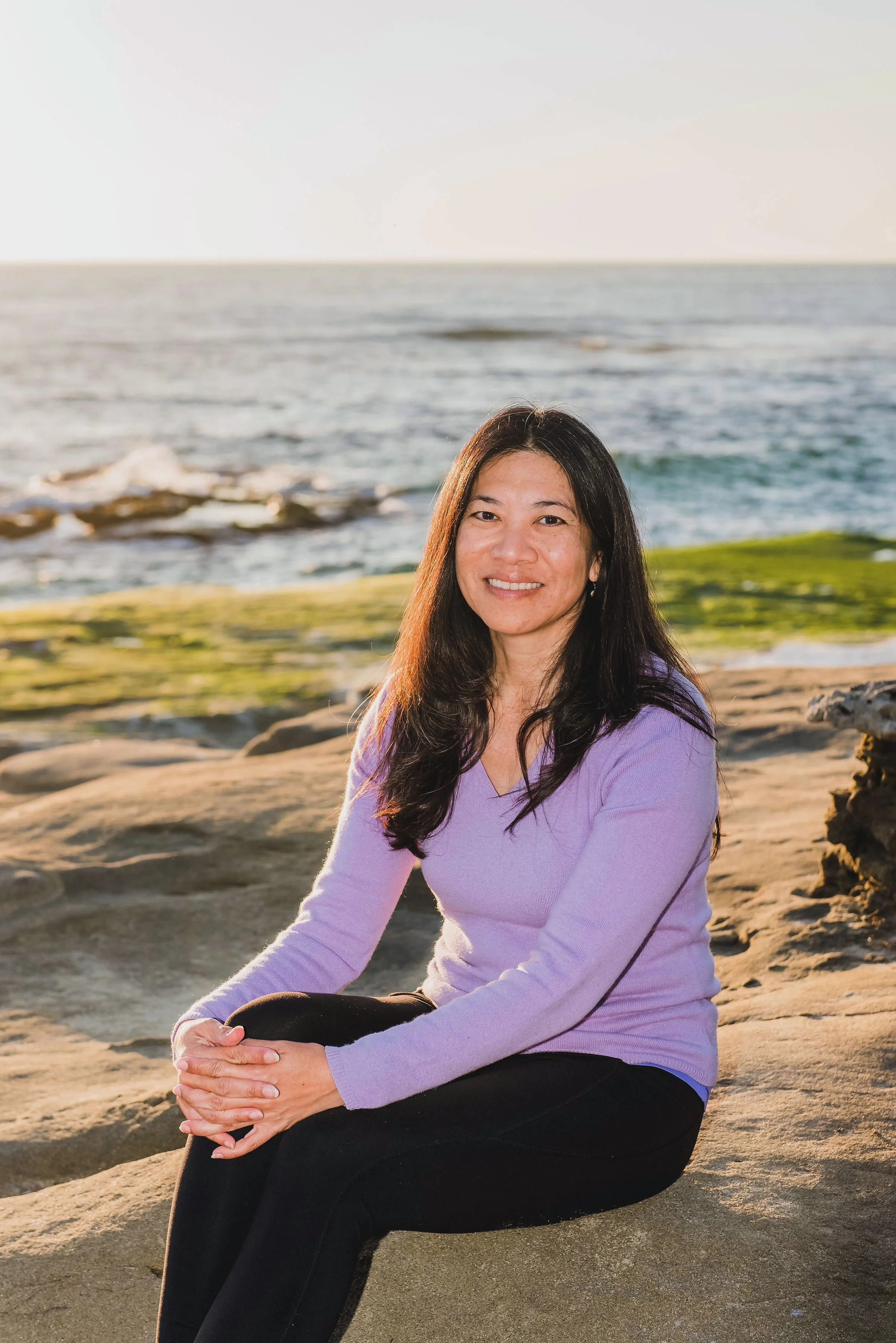A woman sitting on a rock by the beach during sunset, wearing a lavender long sleeve top and black pants, smiling at the camera with the ocean and a clear sky in the background.