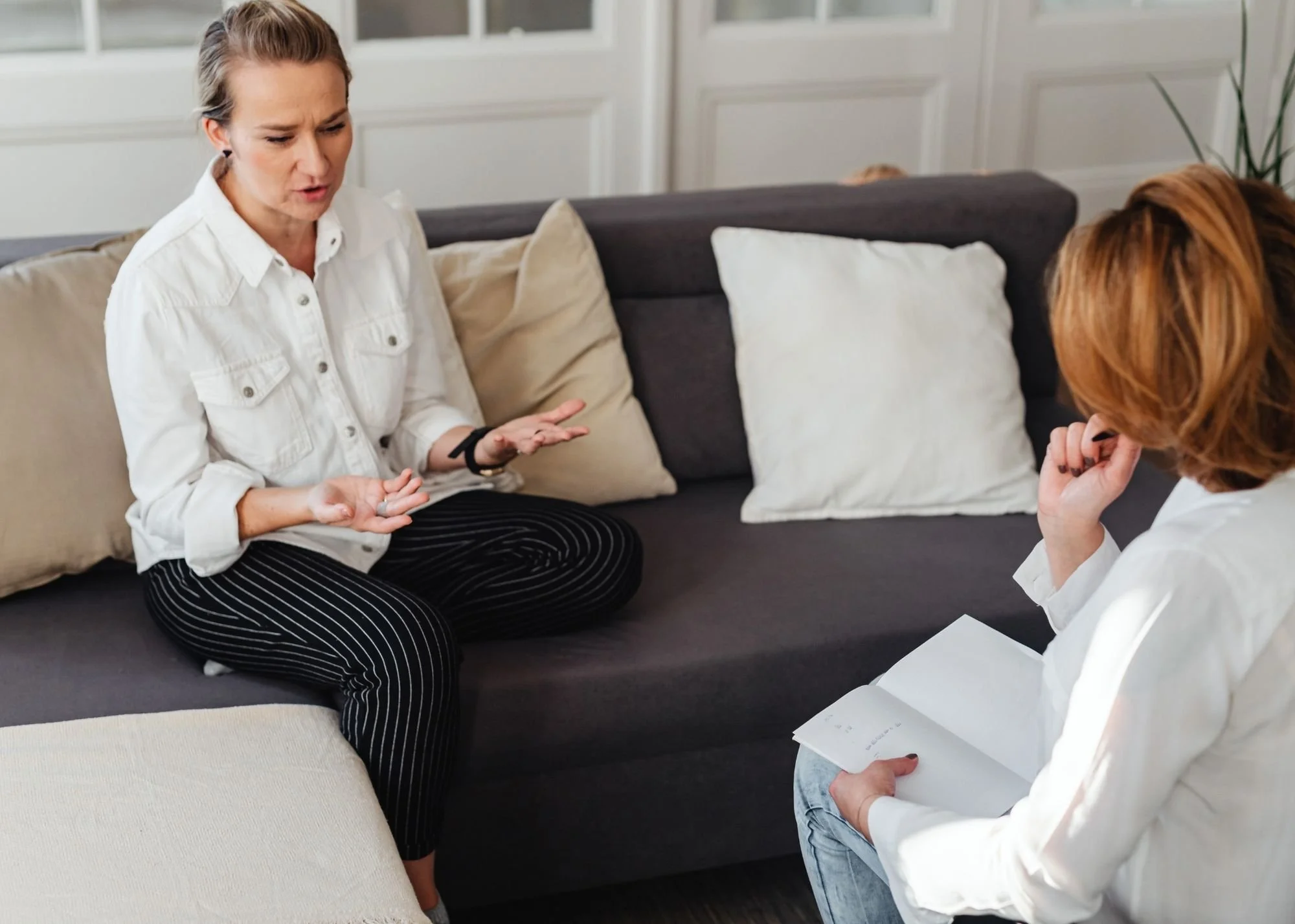 Two women having a conversation in a living room, sitting on a dark-colored sofa with light-colored pillows, with a window and a potted plant in the background.