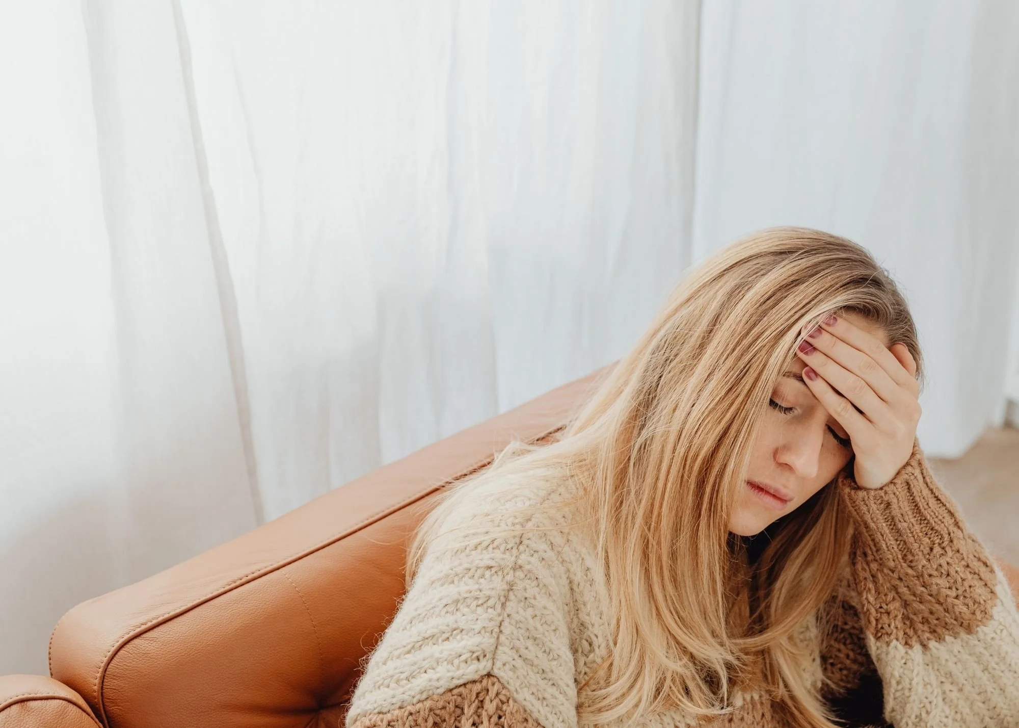 A woman with blonde hair and fair skin sitting on a tan leather couch, touching her forehead with her hand, appearing to be in distress or headache, against a white wall.