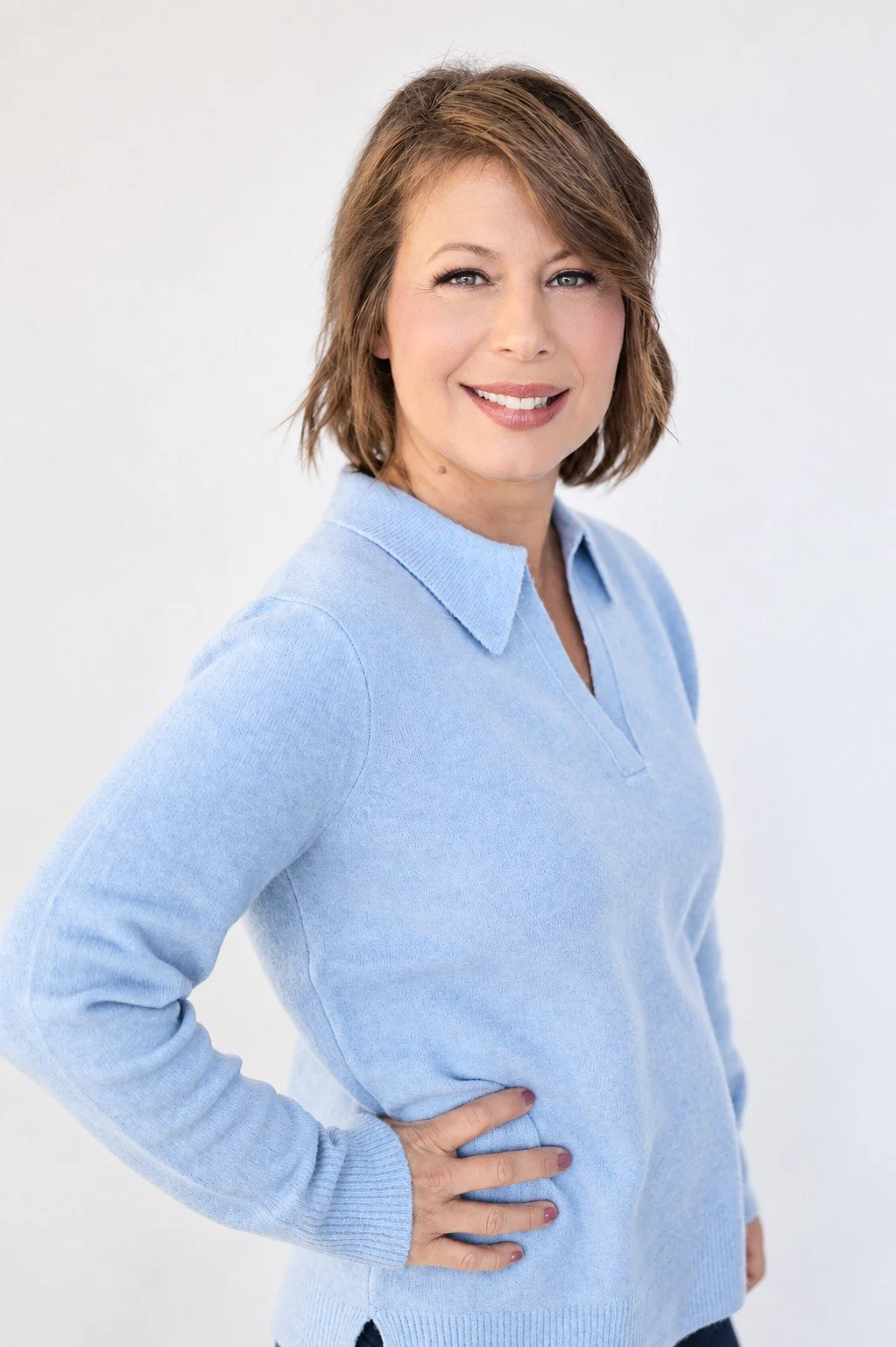 A woman with short brown hair and blue eyes, wearing a light blue sweater, standing with one hand on her hip against a white background, smiling at the camera.