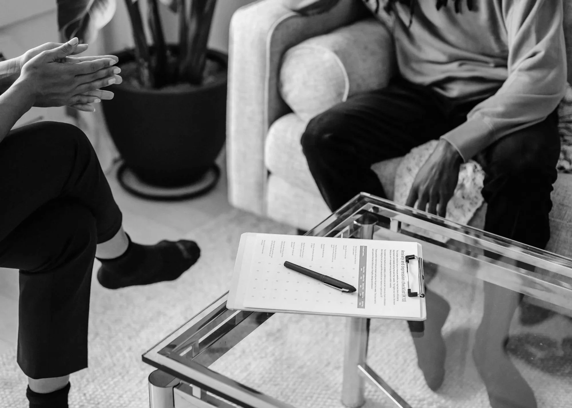 Black and white photo of two people sitting in a waiting area. One person is wearing dark pants and socks, shown with their hands together. The other person is sitting on a cushioned armchair, wearing a light hoodie and dark pants. There is a clipboard on a glass table in front of them, with a pen and a paper that has some printed text.