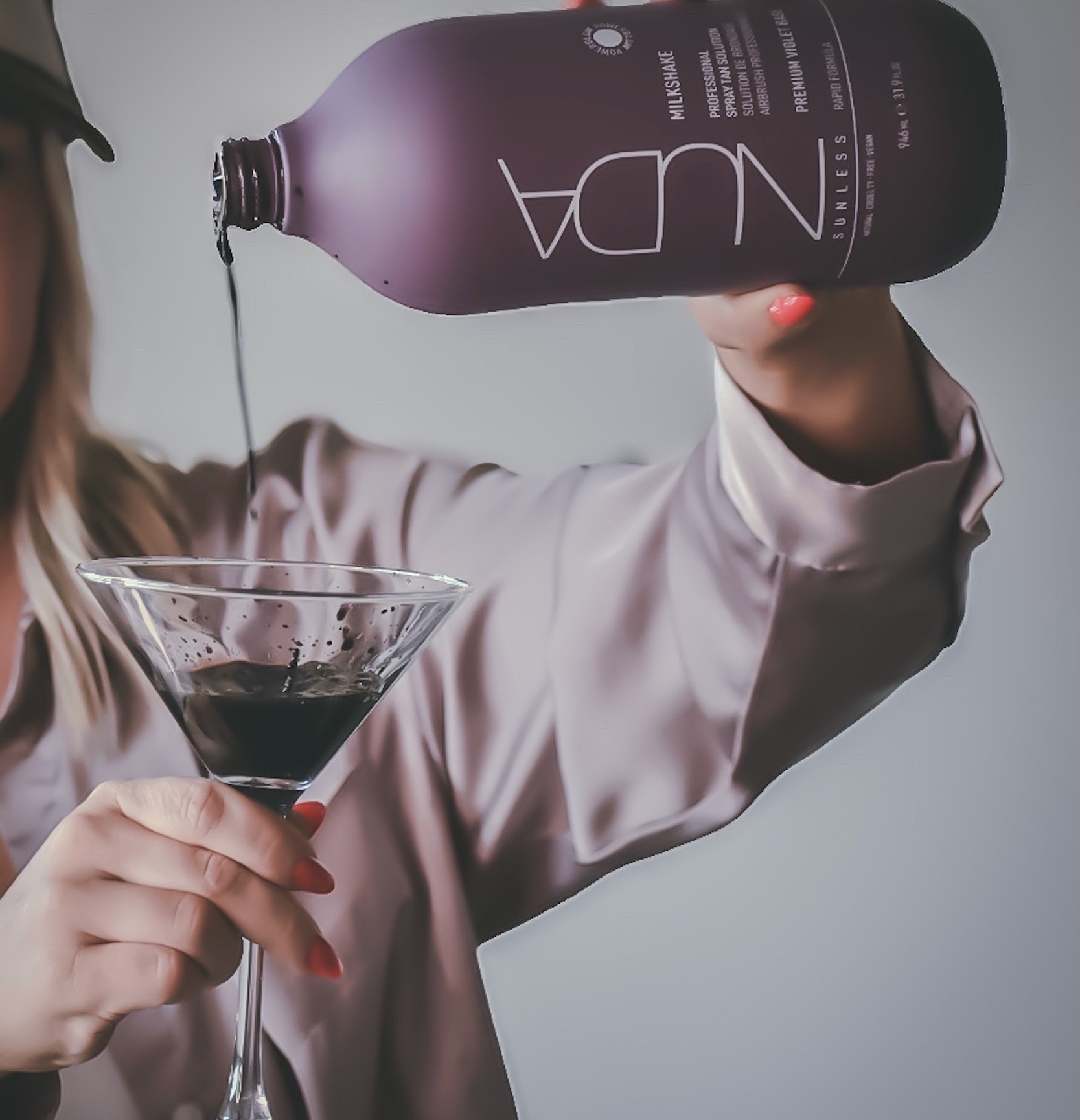 A woman holding a martini glass with a dark liquid, pouring more dark liquid from a NUA Sunless Milkshake bottle.