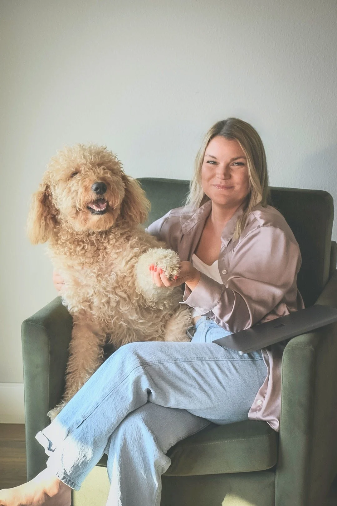 A woman with blonde hair sitting in a green armchair, smiling, holding a large fluffy tan poodle on her lap.