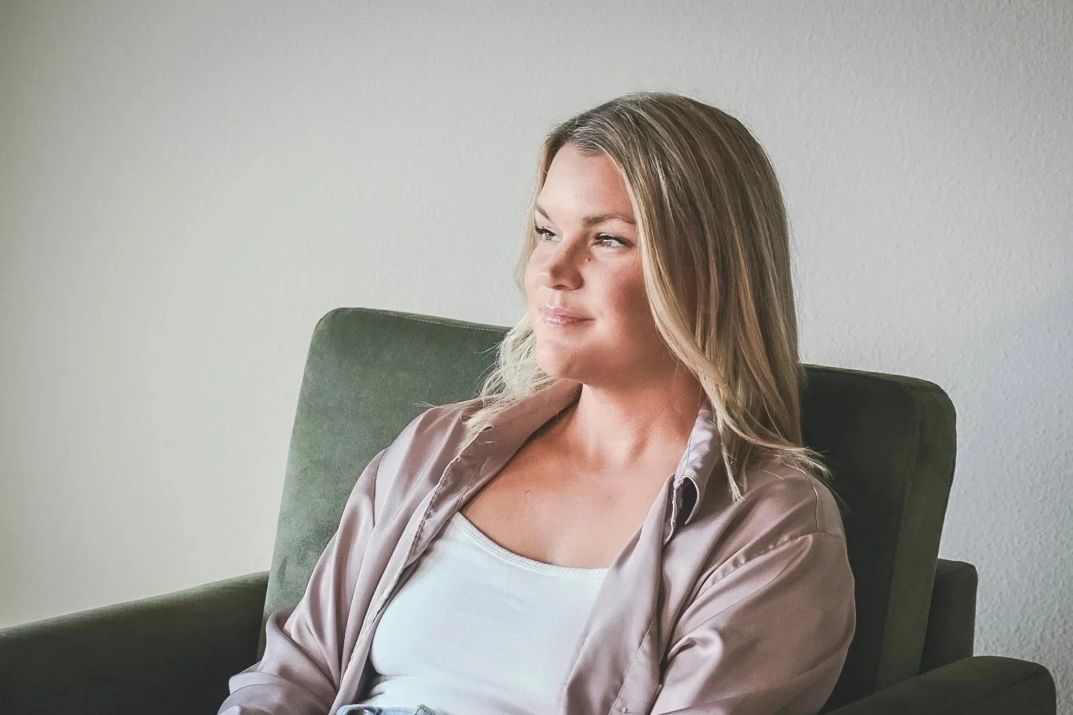 A woman sitting in a green armchair, gazing thoughtfully to the side with a slight smile, wearing a satin beige shirt and a white top, against a plain light-colored wall.
