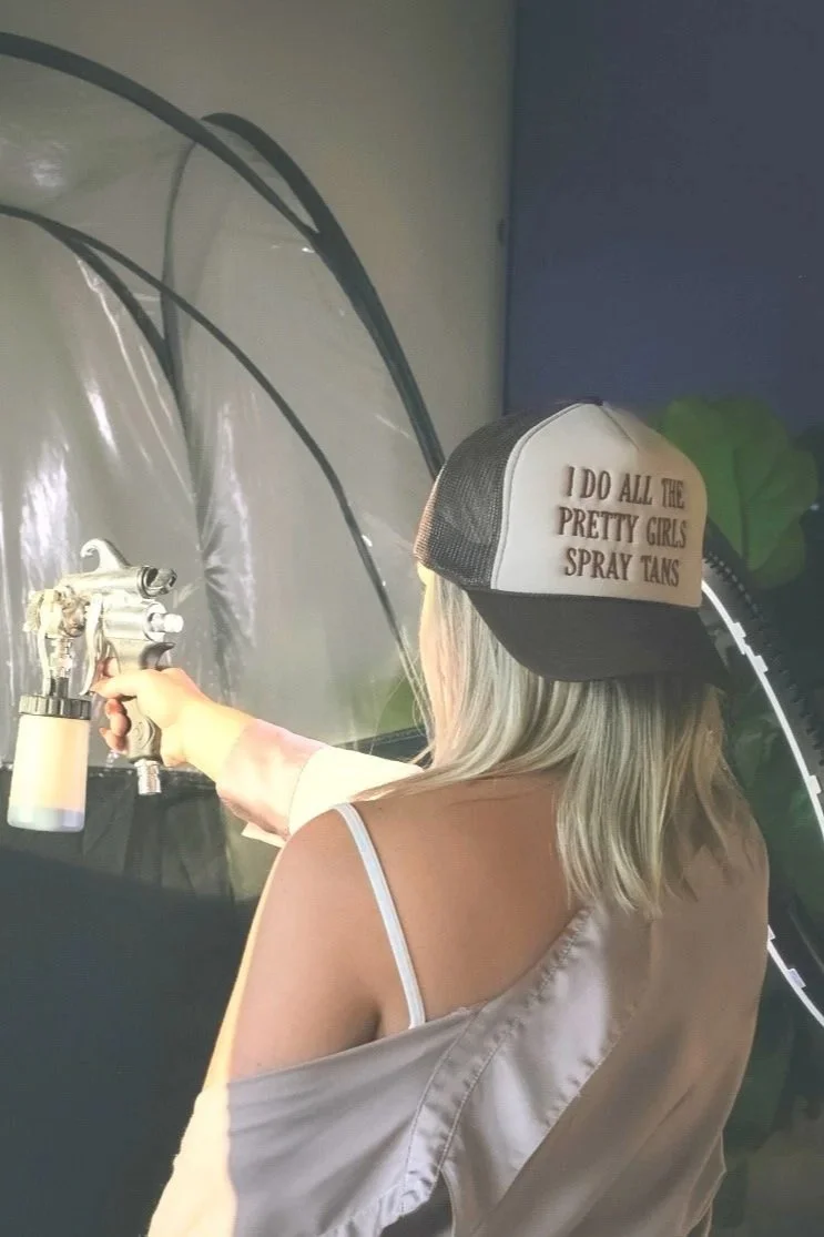 A person with blonde hair wearing a baseball cap that says 'I DO ALL THE PRETTY GIRLS SPRAY TANS', holding a spray tan device, and wearing a beige off-shoulder top with a white strap underneath.