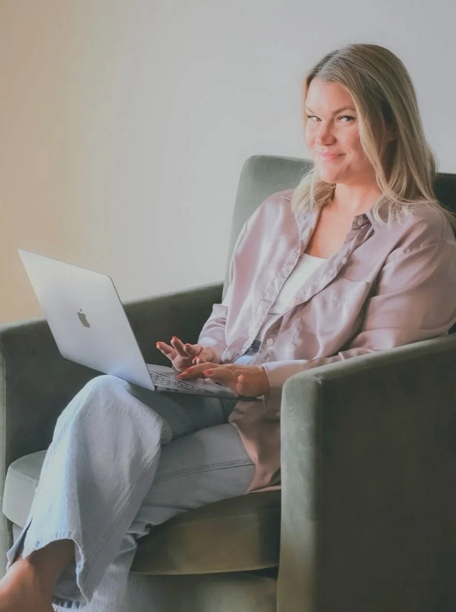 A woman with blonde hair sitting comfortably in a grey armchair, using a silver MacBook laptop, wearing a satin pink shirt and white pants, smiling at the camera.