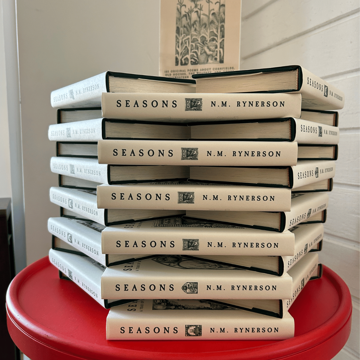 A stack of multiple copies of the book 'Seasons' by N.M. Rynerson on a red round table.