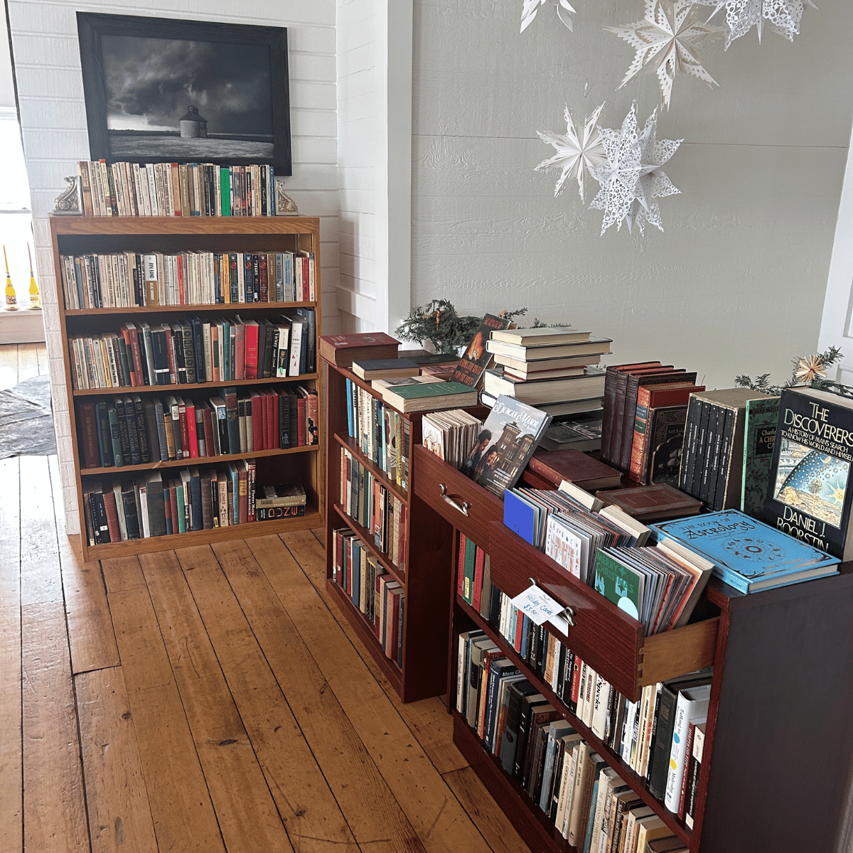 Bookshelves filled with books in a cozy room decorated with white paper snowflakes and a framed black-and-white photograph of a lighthouse, with a wooden floor.