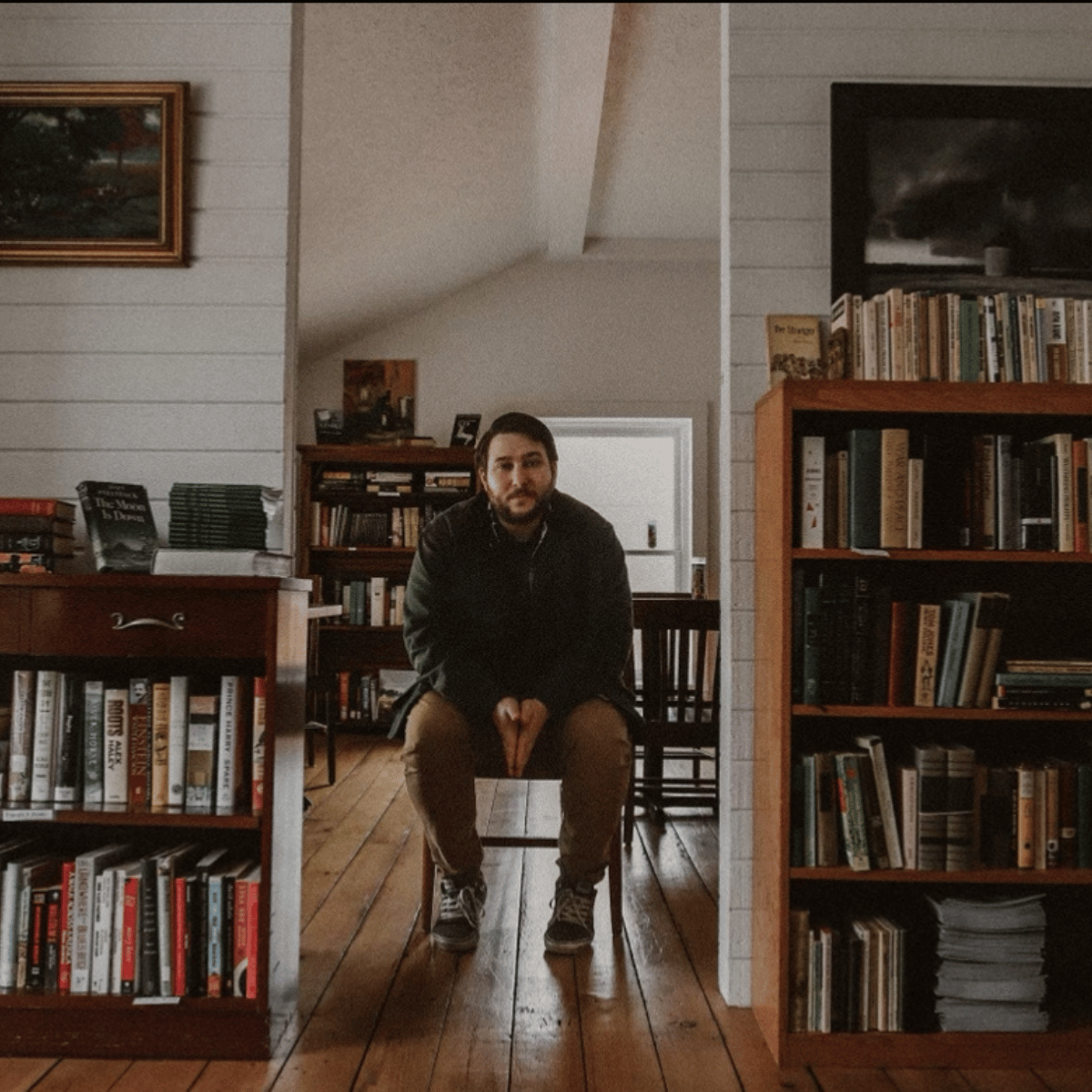 A man sitting on a chair in a cozy, book-filled room with wooden floors and white shiplap walls, looking directly at the camera.