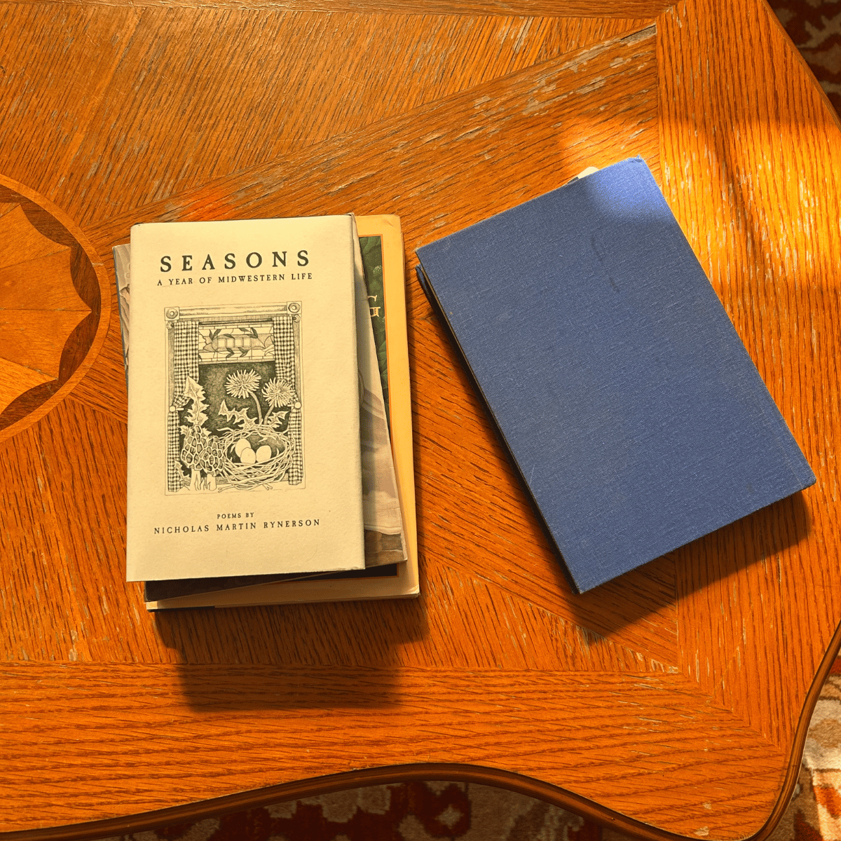 A wooden table holds a small stack of books and a blue notebook. The top book is titled "Seasons: A Year of Midwestern Life" with poems by Nicholas Martin Ryerson, and has a black and white illustration on the cover.