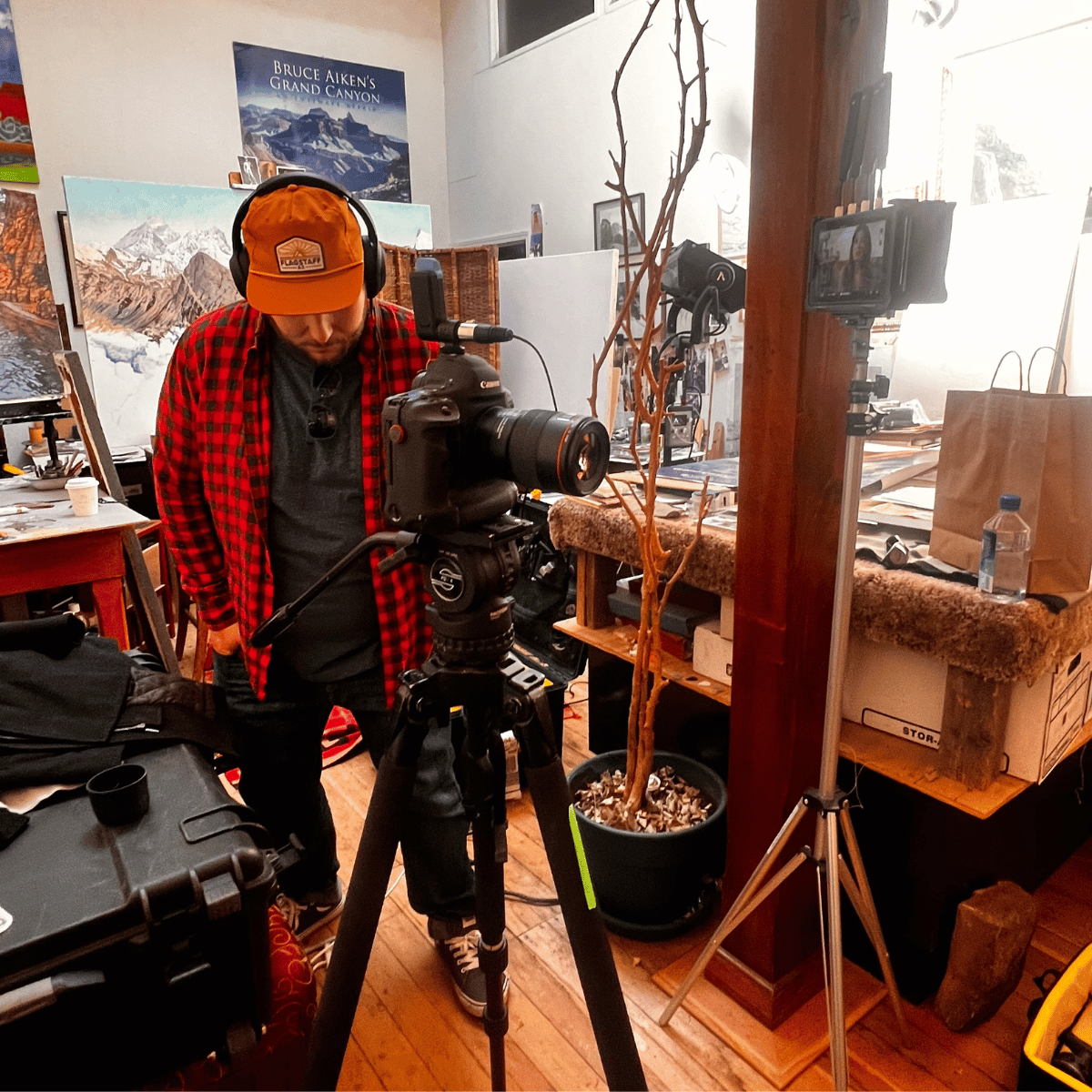 A person wearing a red and black plaid shirt and a yellow hat with headphones, operating a professional camera on a tripod in an art studio, with mountain-themed paintings in the background.