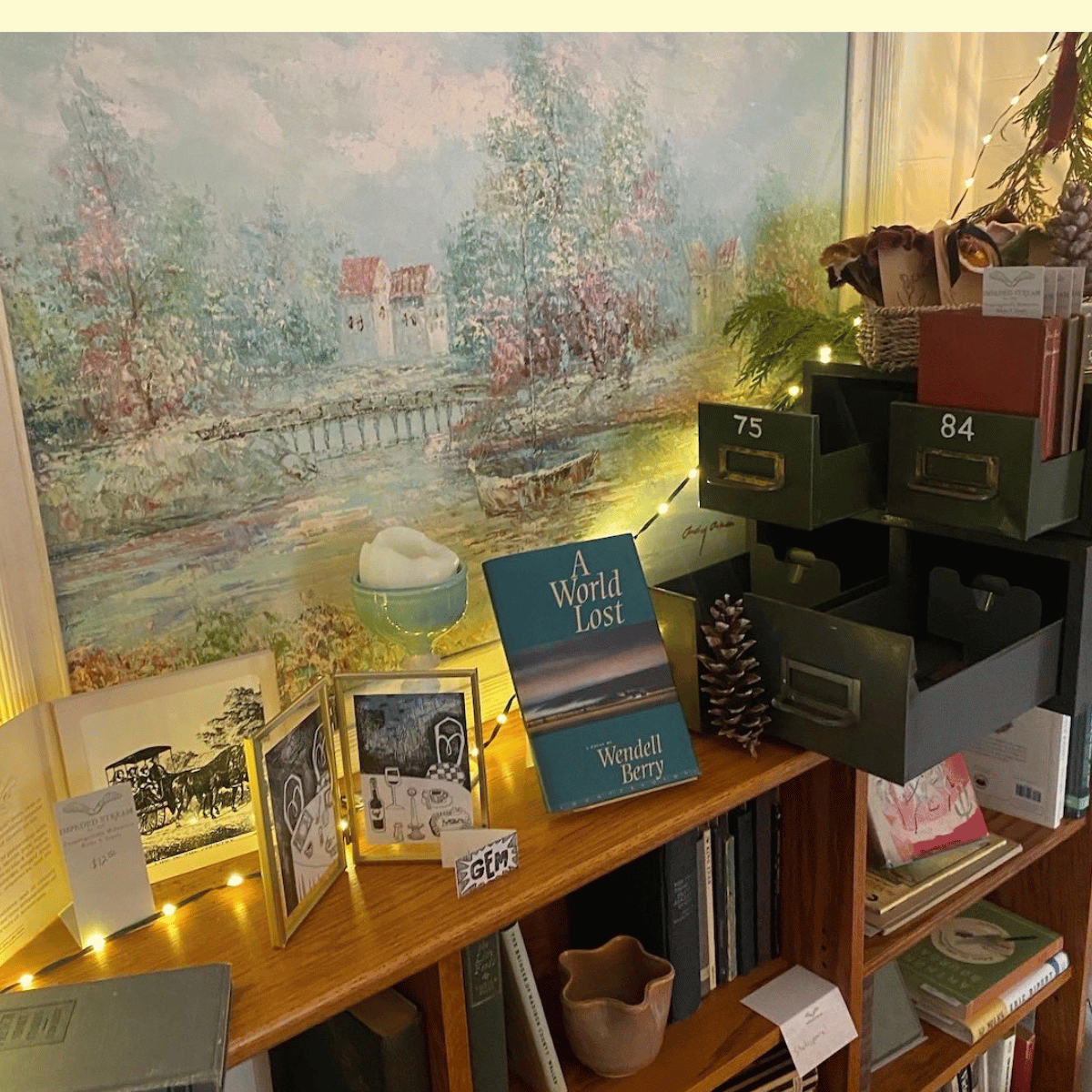 Bookshelf with a painting of a river landscape in the background, a book titled 'A World Lost' by Wendell Berry, framed photos, small decorative items including a pine cone, a bowl with cotton balls, and a string of fairy lights.