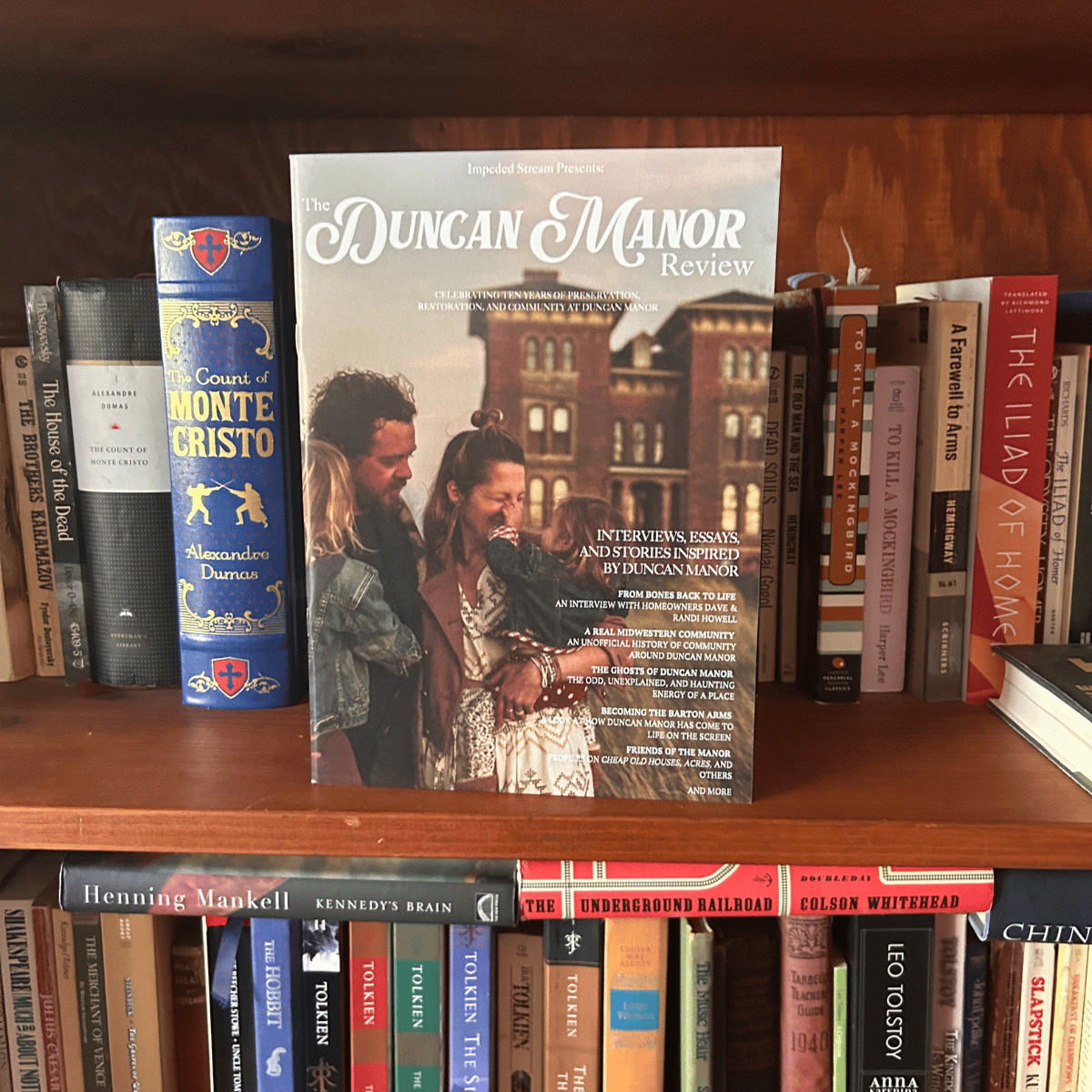 A magazine titled 'Duncan Manor Review' placed on a bookshelf, featuring a cover photo of a woman holding a child, with a man and a woman smiling nearby, and a background of a brick building.