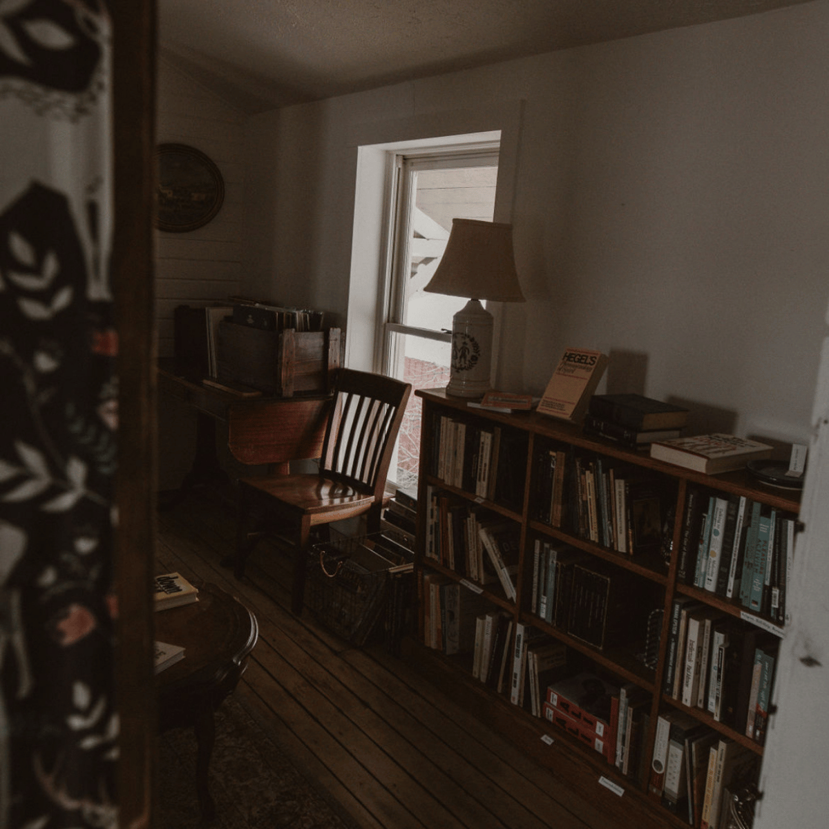 A cozy interior room with a wooden bookshelf filled with books, a wooden chair, a table with books, a large table lamp, and a window letting in natural light.