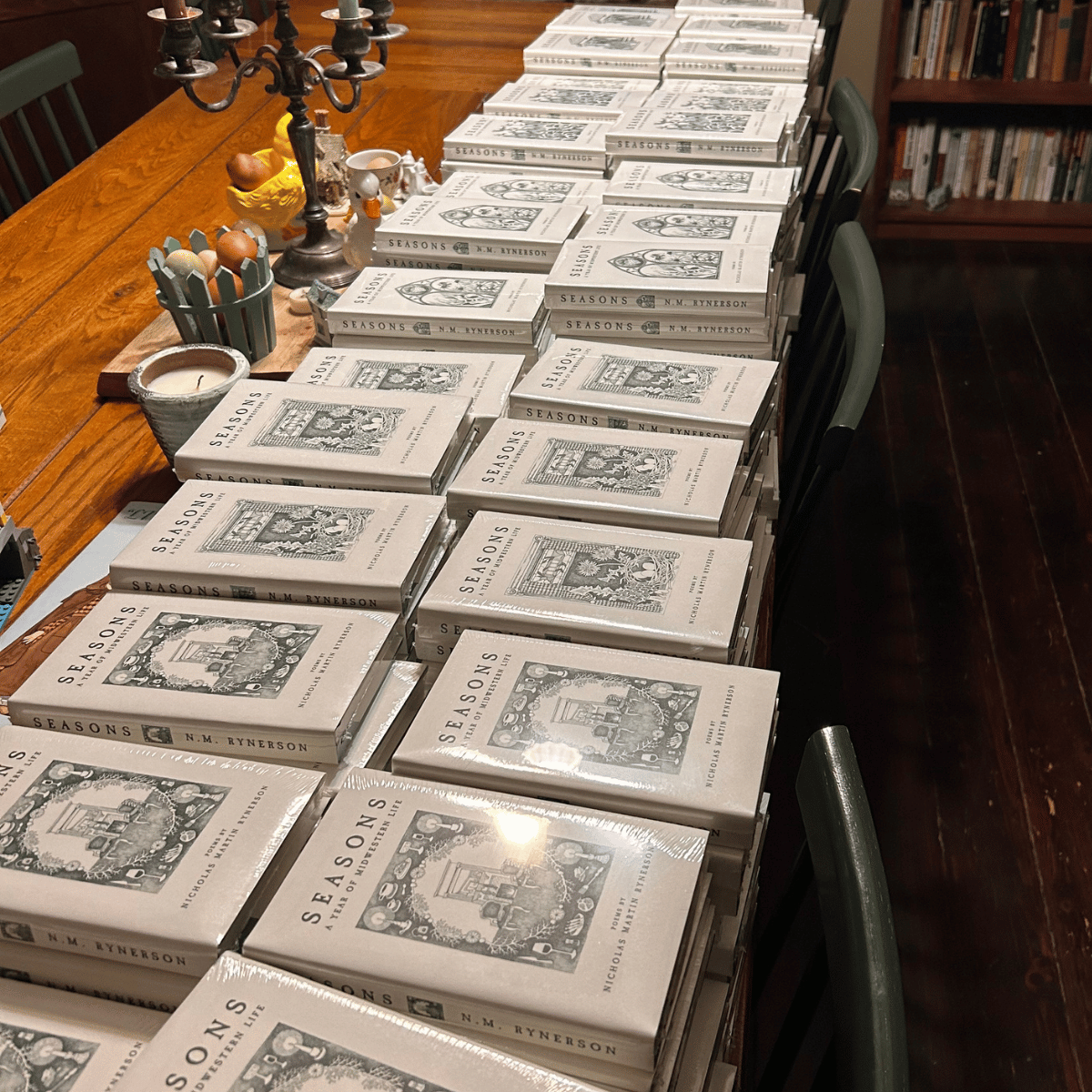 A table covered with multiple copies of the book 'Seasons' by N. M. Ryerson, along with decorative items like a candelabra, eggs, and a small teapot.