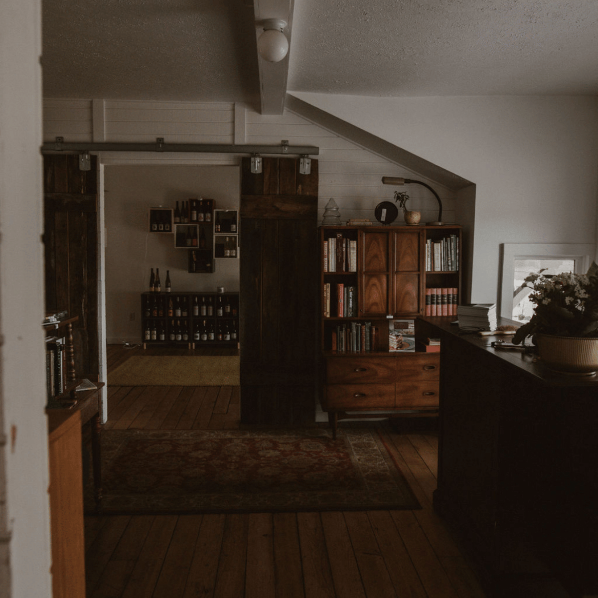 A cozy living room with wooden furnishings, bookshelves filled with books, and a dark wood sideboard, illuminated by natural light from a nearby window.