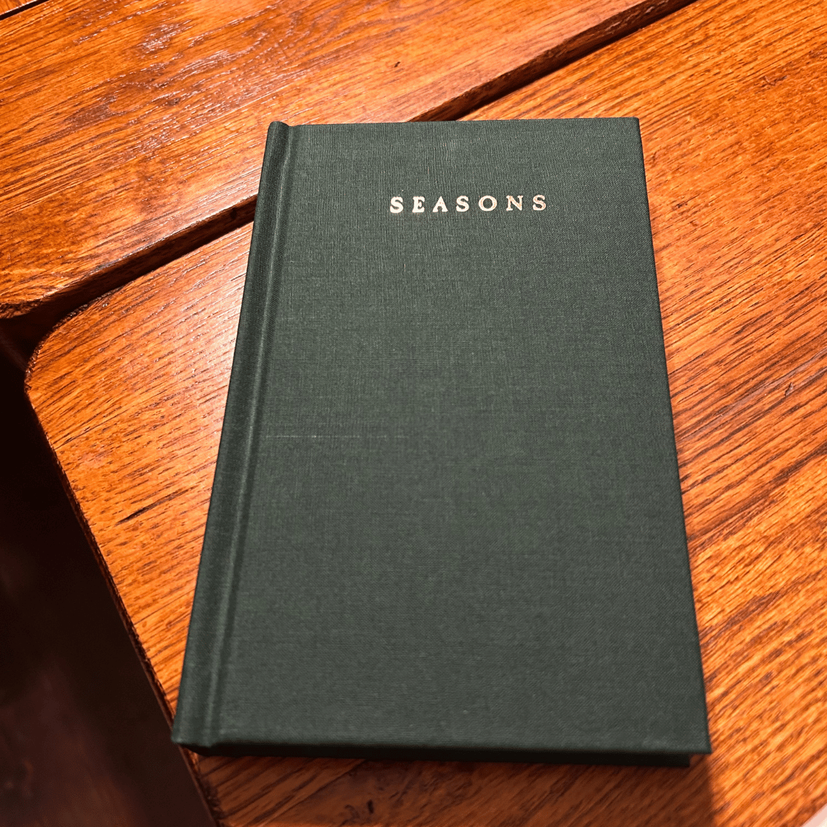 A green hardcover book titled 'Seasons' resting on a wooden table.