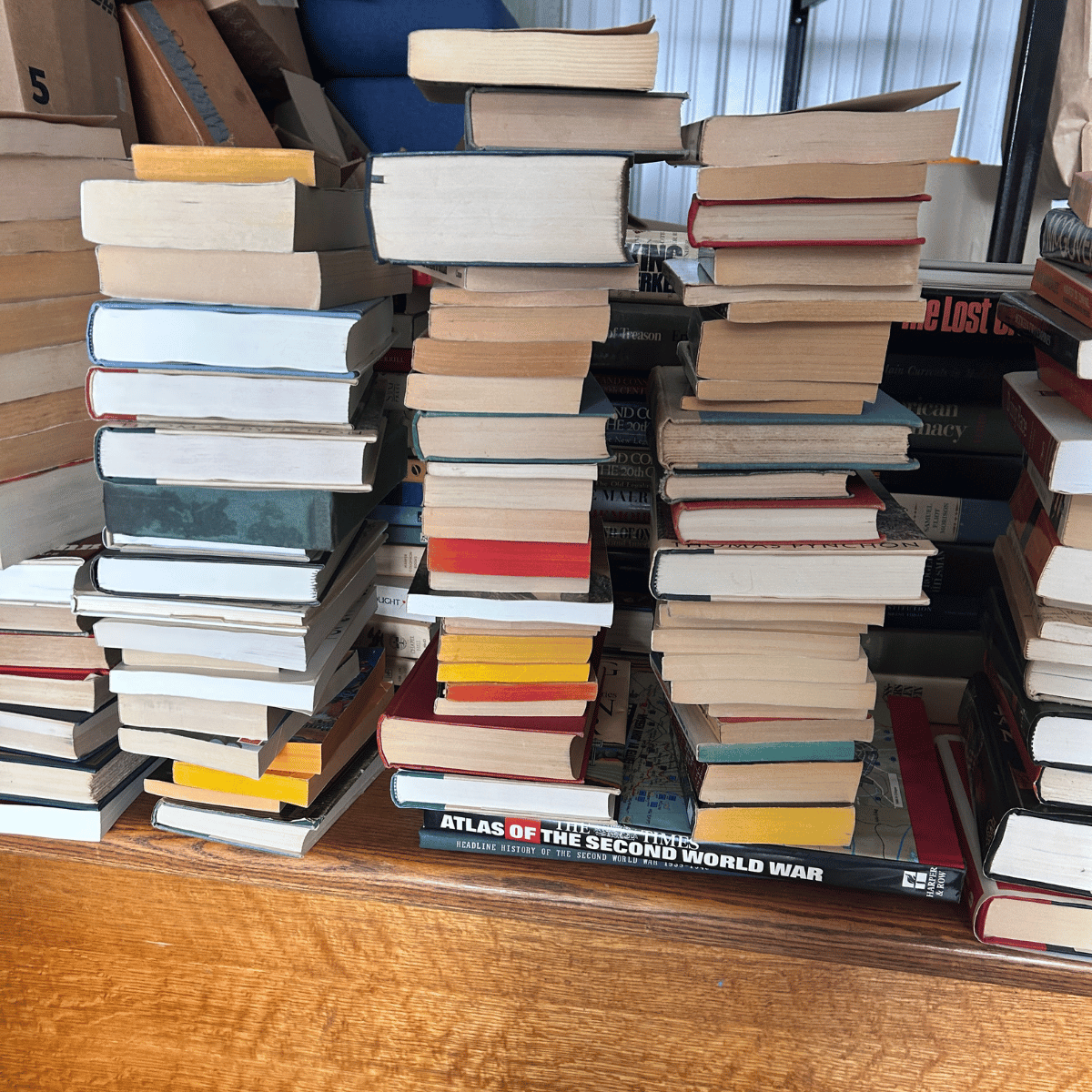 A large stack of books on a wooden table, with some books placed on top of each other and others leaning. The books vary in size, color, and condition, with some titles partially visible.