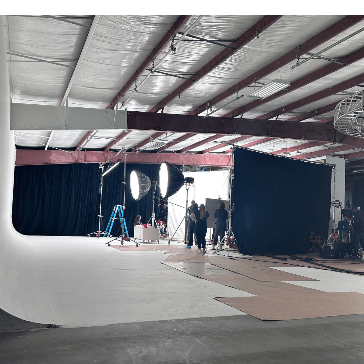 Behind the scenes of a professional photography or video shoot, featuring black curtains, large studio lights, crew members, and equipment.