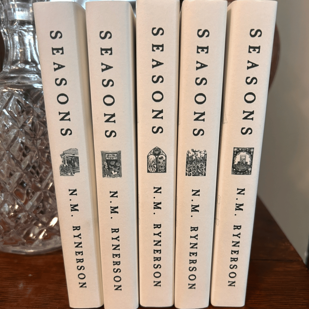 A collection of five books titled 'Seasons' by N. M. Rynerson, standing upright on a wooden surface with a glass decorative jar to the left.