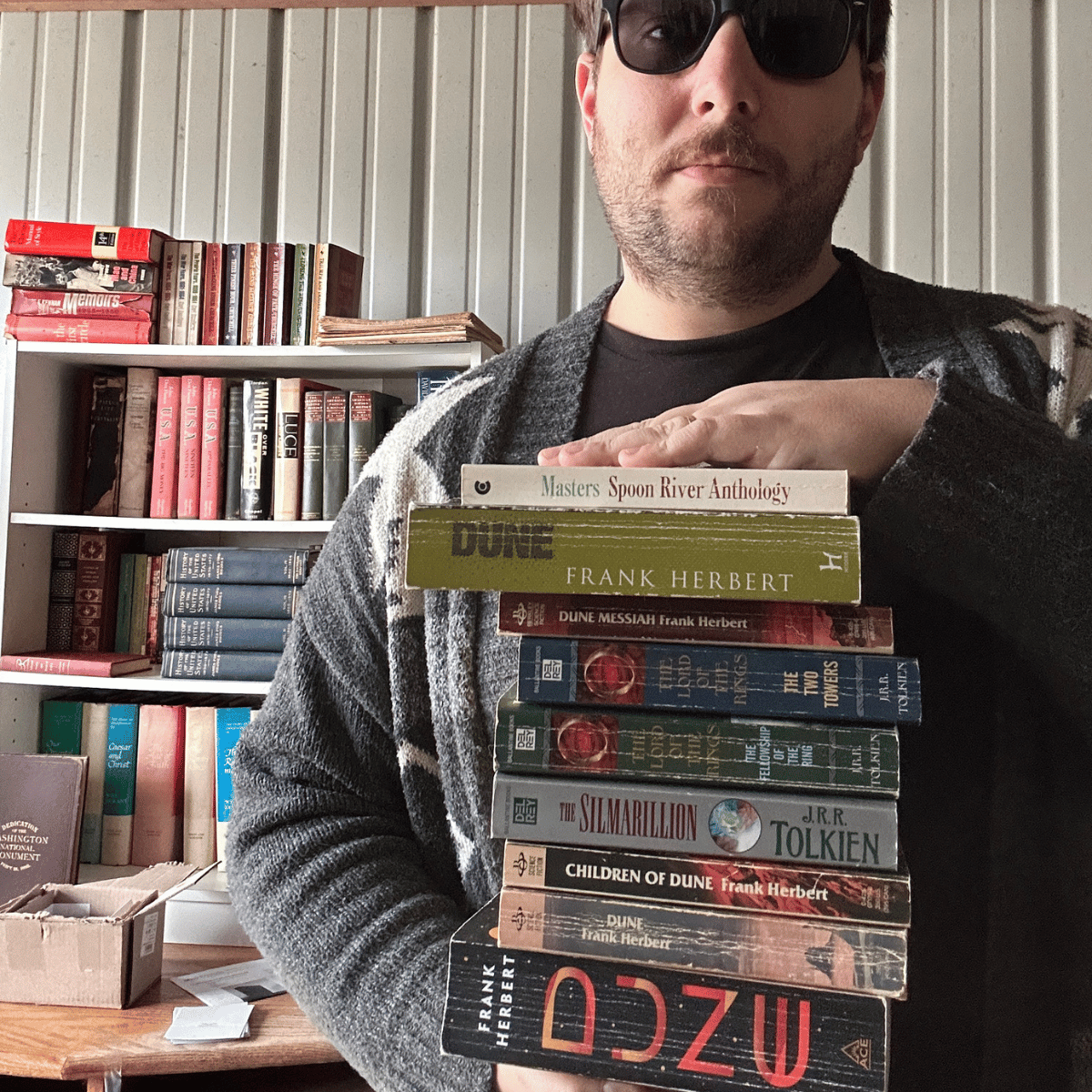 A person with dark sunglasses and a beard holding a stack of classic science fiction books, including titles by Frank Herbert and J.R.R. Tolkien, standing in front of a bookshelf filled with various books.