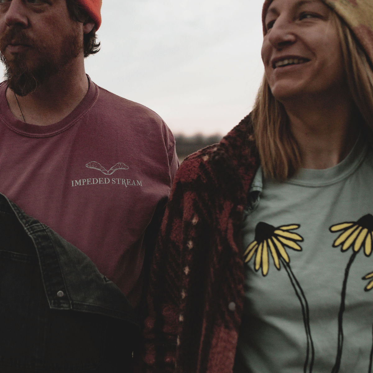 Close-up of two people walking outdoors, wearing casual clothing, with one wearing a shirt that says 'Impeded Stream' and a graphic of leaves, and the other wearing a shirt with a flower design under a plaid jacket, during cloudy weather.