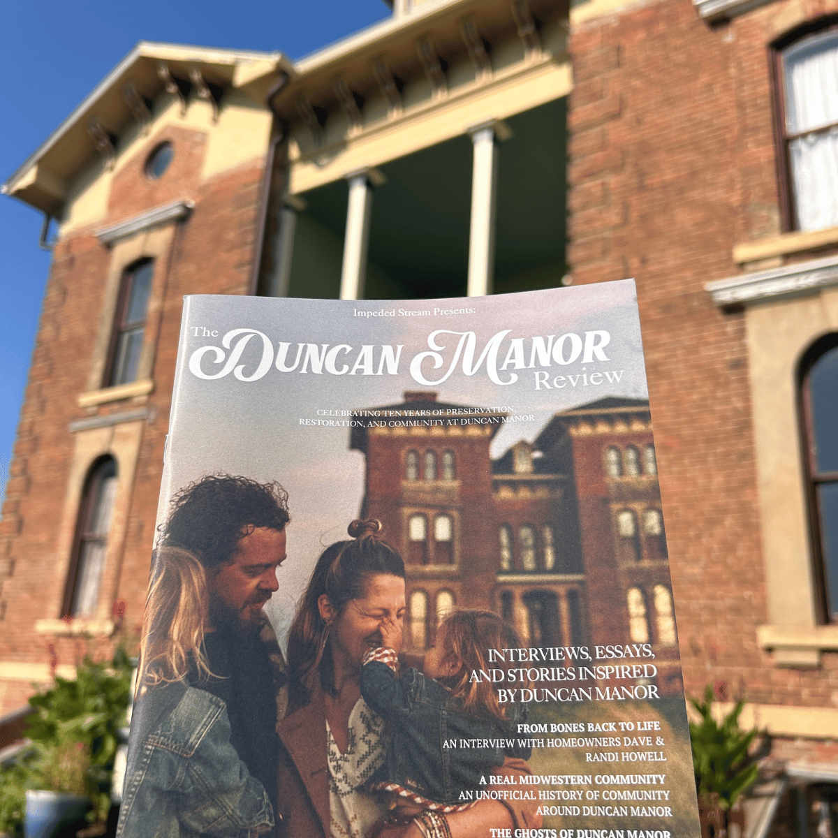 A person holding a magazine titled "The Duncan Manor Review" in front of a historic brick building with large windows and a clear blue sky.